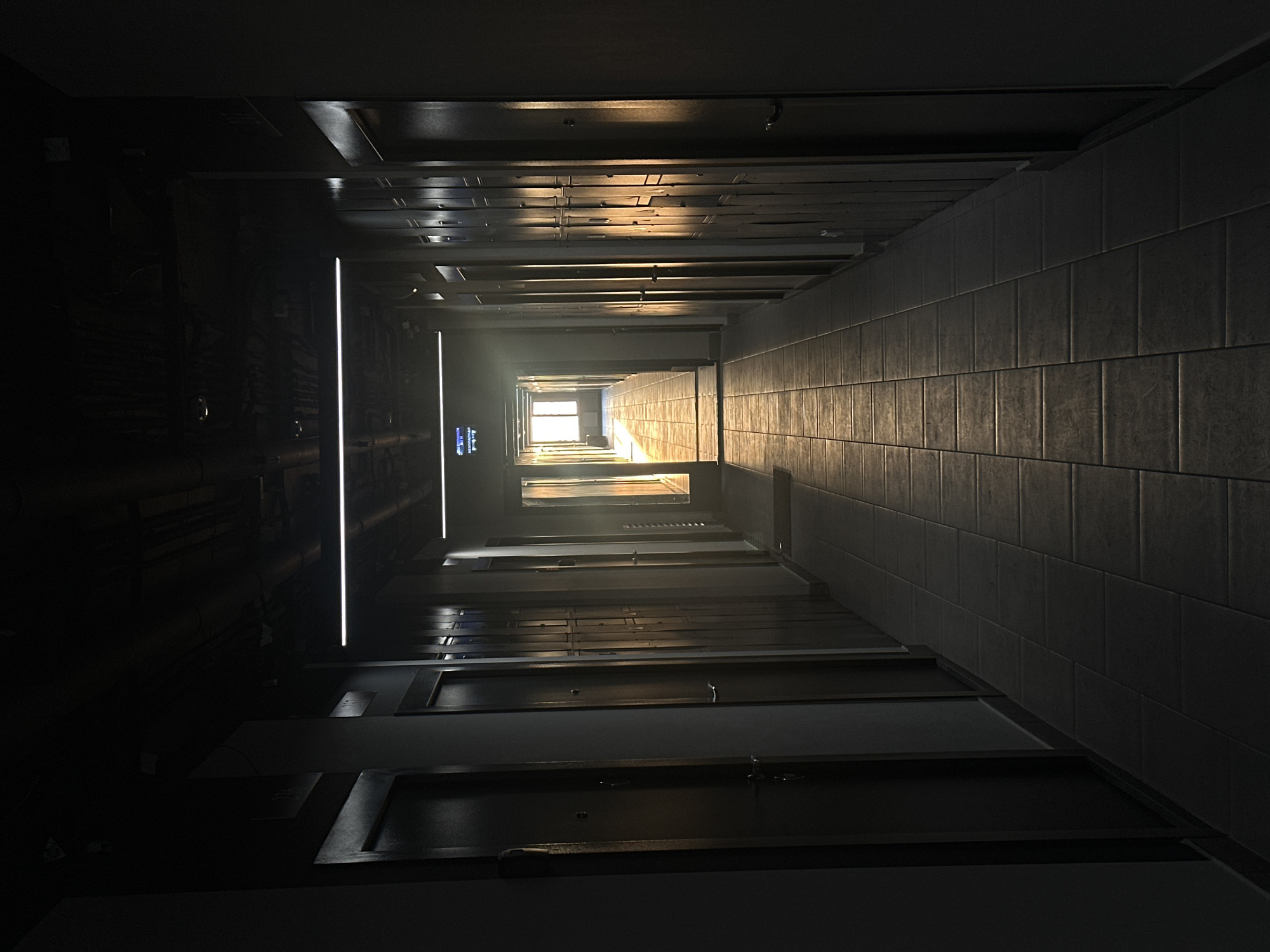 The image depicts an interior hallway with tiled walls and multiple doors. The hallway recedes into the distance with a light source visible at the far end. The lighting creates a dramatic effect, with strong contrasts between light and shadow. There are linear light fixtures along the left side of the hallway. The location is the city of Khimki, Russia, but there is no information to determine that is a part of the image.