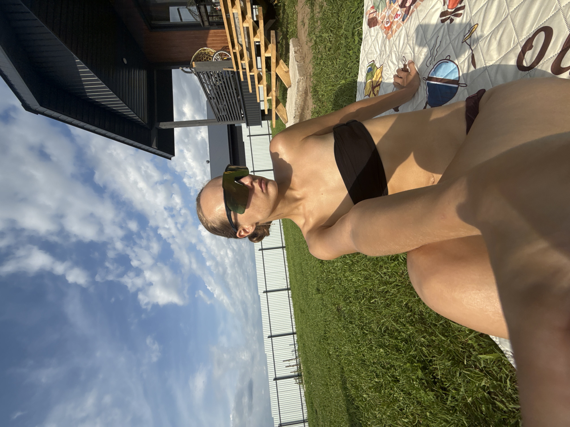 In the city of Podshivalovo, Russia, a woman is lying on a blanket on the grass, facing the camera and wearing a black bikini top and sunglasses. Her hair is pulled back, and she has a silver earring in her visible ear. Behind her is a white fence and a wooden house with a dark roof and a wooden staircase. The sky above is blue with scattered white clouds, and the sun is shining, casting shadows on the grass and the woman.