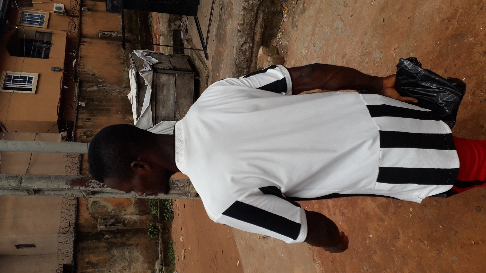A person wearing a white shirt with black stripes on the sleeves and back is holding a black plastic bag. The person is standing on a reddish-brown surface. The background shows buildings with orange walls and barbed wire, along with a makeshift structure made of wood and white tarp. The scene appears to be set in Nkpor, Nigeria.