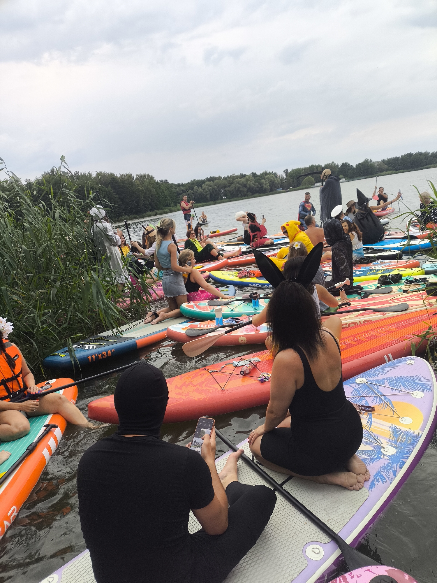 A group of people in various costumes are gathered on paddleboards on a body of water. The people are dressed as characters such as Superman, a scarecrow, a witch, and a bunny. Some are holding paddles, while others are interacting with each other or looking at their phones. The environment appears to be a lake or calm river with trees and foliage lining the shore in the background. The sky is overcast. The scene is likely a recreational event or party on the water.