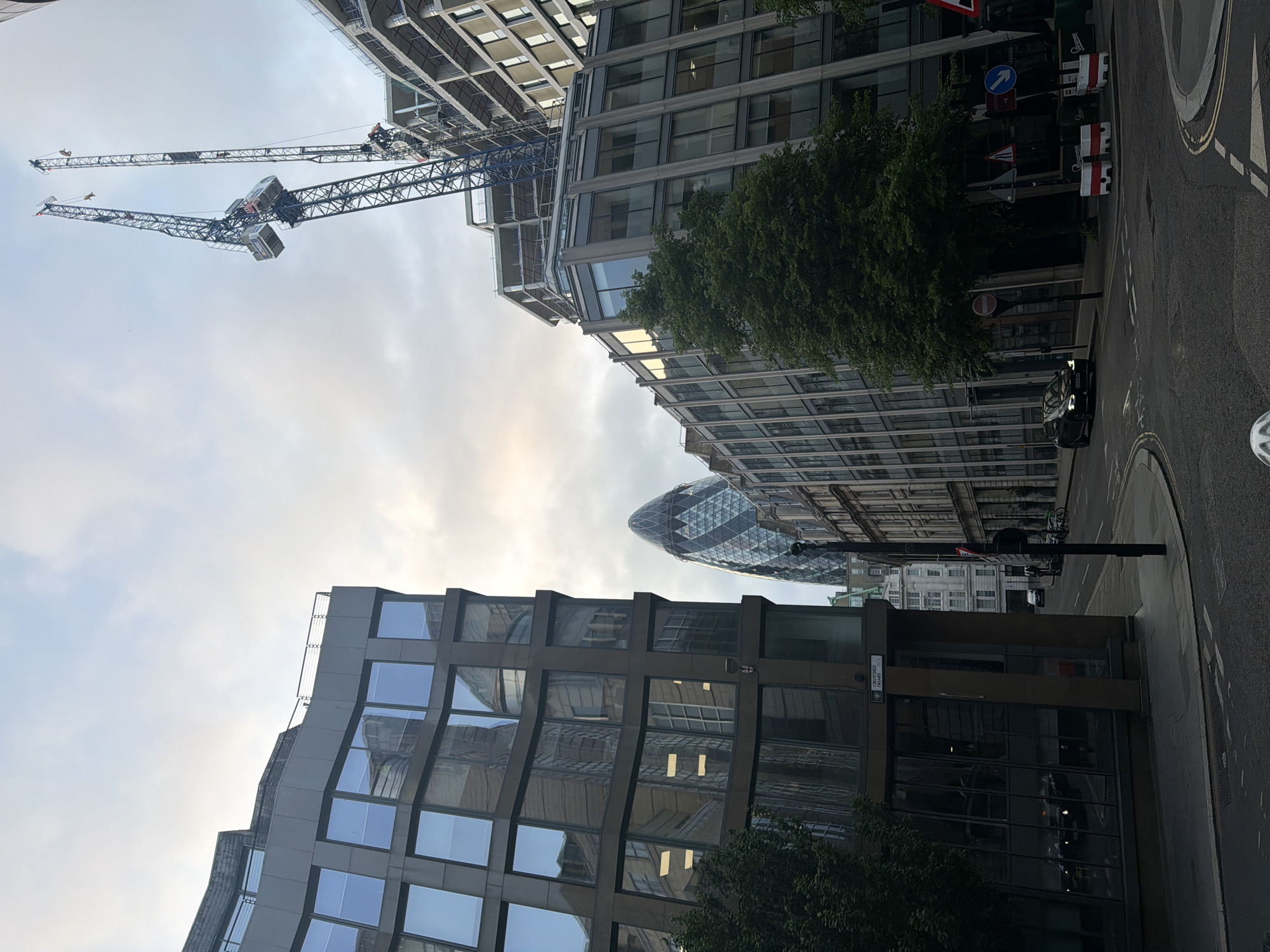 The image captures a street view in London, United Kingdom, featuring modern architecture and construction. In the background, the distinctive shape of "The Gherkin" building (30 St Mary Axe) is visible. Construction cranes dominate the upper portion of the image, juxtaposed against a building under construction. Various office buildings with glass facades are interspersed with green trees. A dark vehicle is on the street, and street signs are visible on the sidewalk.