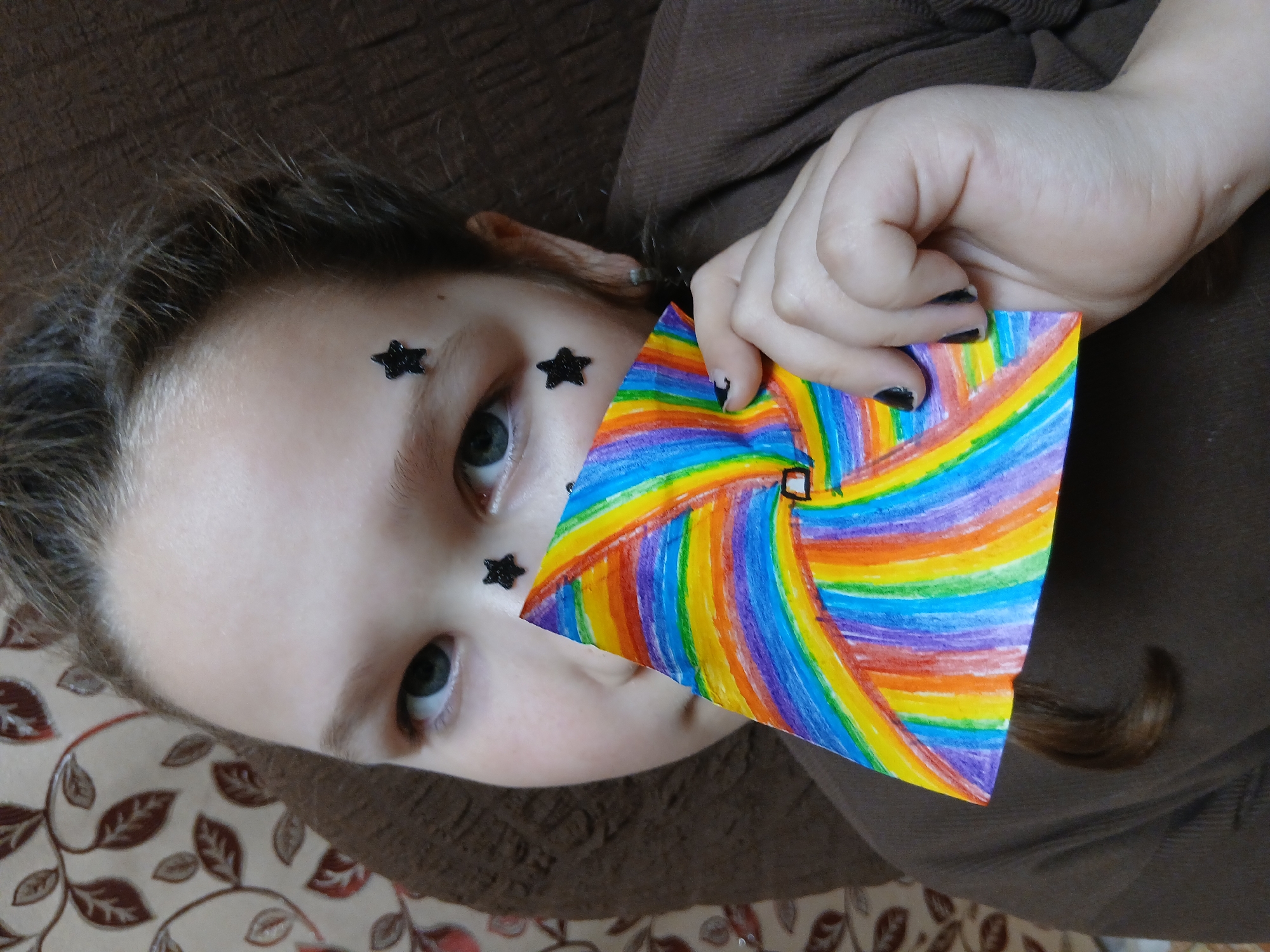 A young person with star-shaped stickers on their face is holding a square piece of paper with rainbow-colored concentric lines. The person is lying down, with a brown shirt visible and a patterned surface underneath. The location is not evident from the image.