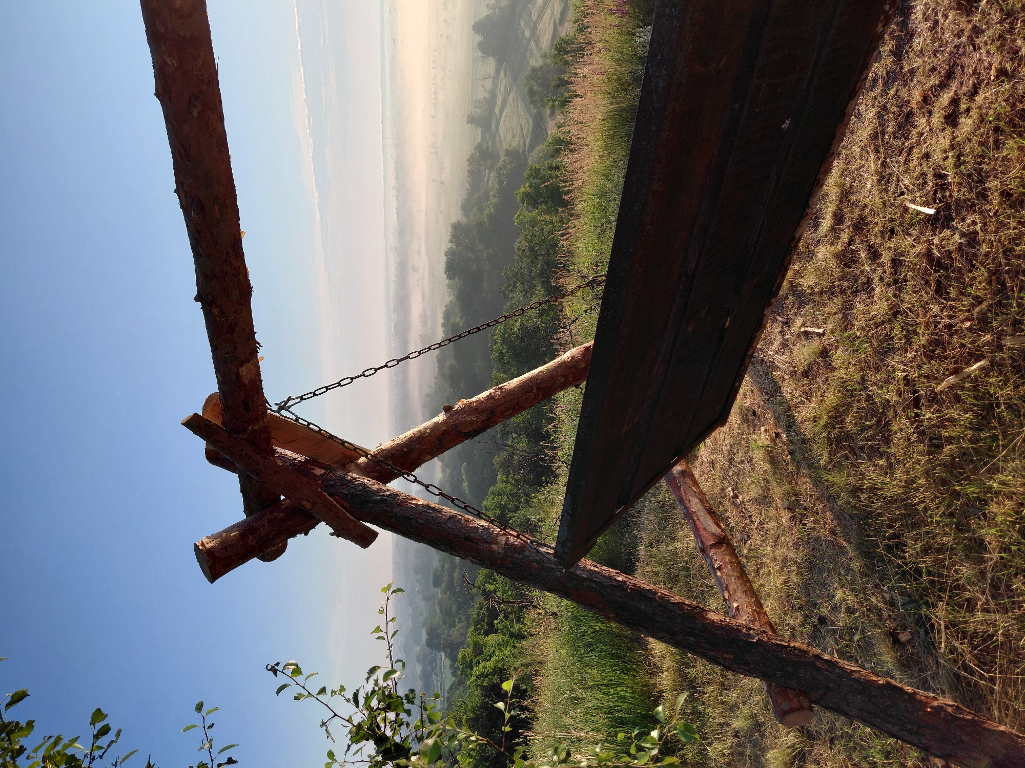 In Ustyvytsia, Ukraine, a wooden swing frame stands on a grassy knoll overlooking a misty valley. The swing itself, a dark wooden plank suspended by chains, is partially visible in the foreground. The sky is a clear blue with a faint haze in the distance, suggesting early morning or late evening light. Trees and rolling hills fill the landscape, partially obscured by the morning mist. The ground around the swing is covered in dry grass, with a few scattered twigs. A branch with green leaves hangs from the upper left corner of the frame.