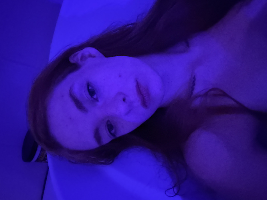 A young adult female with reddish-brown hair is depicted in a close-up, chest-up view, submerged or reclining within a white, tub-like structure. The scene is illuminated by a predominant artificial blue-purple light, casting an even hue over the subject and surroundings. The female's gaze is directed forward, towards the camera, with a neutral to slightly pensive expression. Her skin exhibits minor imperfections, including a noticeable mole on her right cheek (viewer's left) and small blemishes on her forehead. A dark, wavy tattoo is visible on her upper left chest/collarbone area (viewer's right). Her bare shoulders and upper torso are visible above what appears to be water or the tub's edge. In the upper left background, a dark, cylindrical container or object is faintly discernible against the blue-lit wall. The location of this scene is the city of Khimki, Russia.
