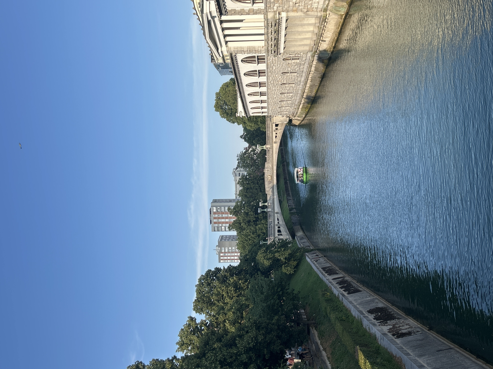 This is a landscape photograph of a canal with a boat on it, taken on a clear, sunny day. On the right side of the canal is a long, multi-story building with classical architectural elements, including columns and arched windows. The building's facade is made of stone. On the left side of the canal is a grassy embankment with trees and some people walking along a pathway. In the background, there is a bridge arching over the canal, and behind that, several modern apartment buildings are visible. The canal itself is a deep blue-green color, reflecting the sky and the surrounding structures. A small, green boat with passengers is moving along the canal towards the bridge. The sky is a clear, bright blue with a faint contrail from an airplane visible in the upper right quadrant. The scene appears to be an urban environment with a mix of historical and modern architecture.