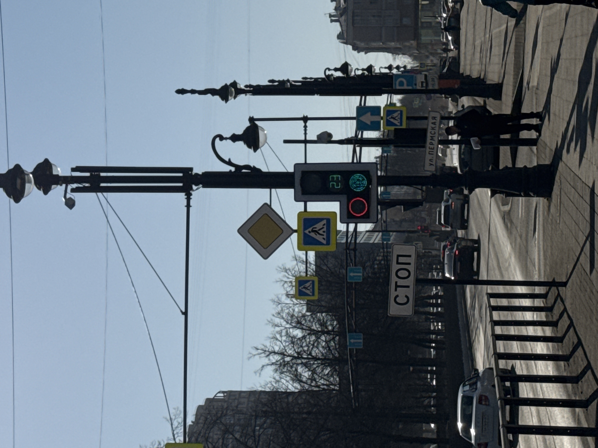 Here's a description of the image:
The image captures a street scene in Perm, Russia, specifically at the intersection of ulitsa Permskaya. The view is from the sidewalk looking out towards the street. Several street signs are visible, including pedestrian crossing signs, a 'Stop' sign, and a sign indicating 'ul. Permskaya'. A traffic light is shown with a countdown timer and a green light signaling that it is safe to cross the street.
Several cars are on the road. There are lamp posts. A pedestrian is also walking on the sidewalk. The buildings line the street. The sky is blue and appears to be sunny.