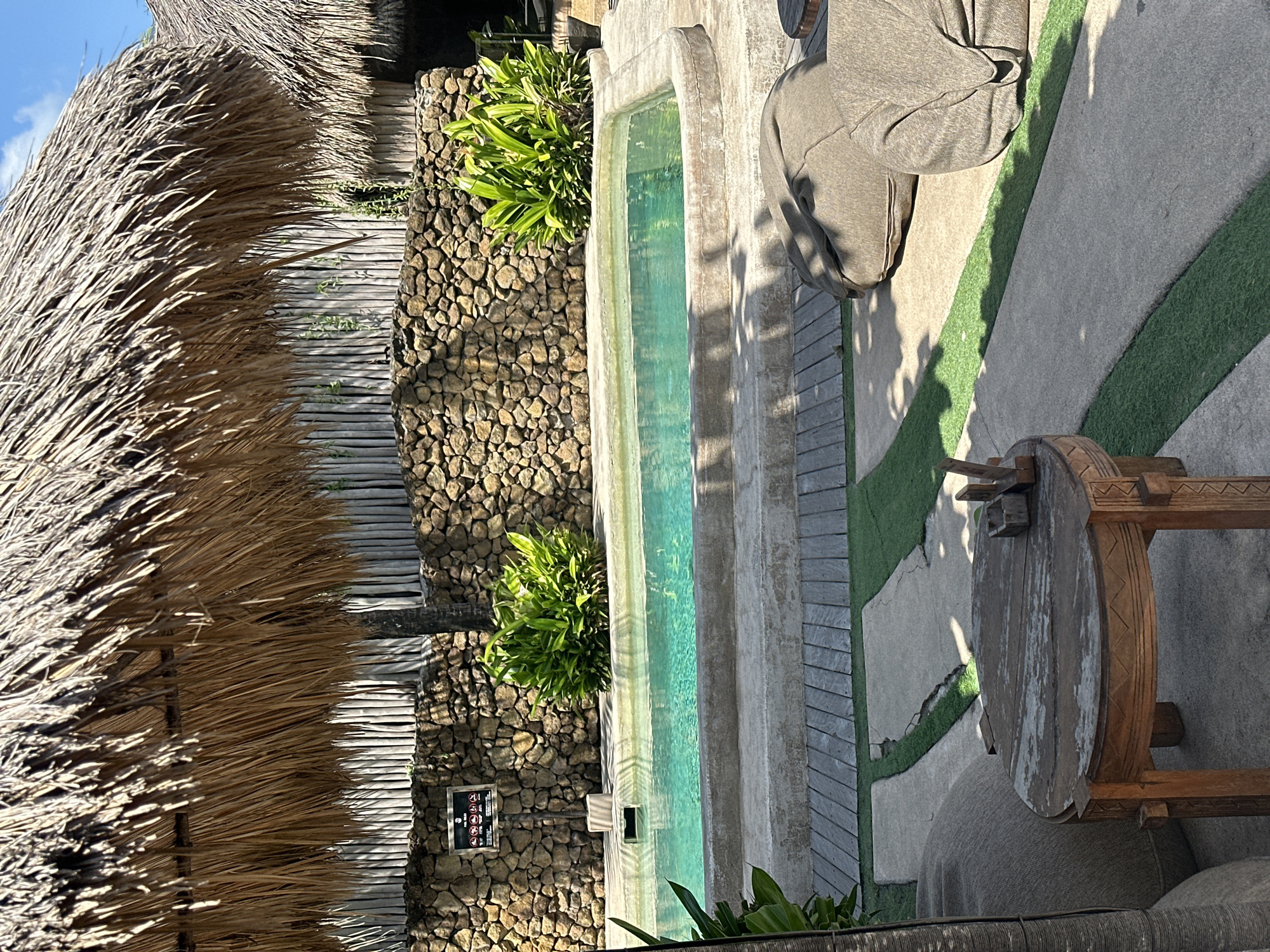 The image shows an outdoor seating area next to a swimming pool at a resort in Kuta, Indonesia. A thatched roof covers part of the scene. A stone wall with wooden fencing behind it forms the backdrop. Lush green plants are visible around the pool. Beanbag chairs and a wooden table with a small wooden organizer are placed on the patio. The swimming pool is filled with clear blue water.