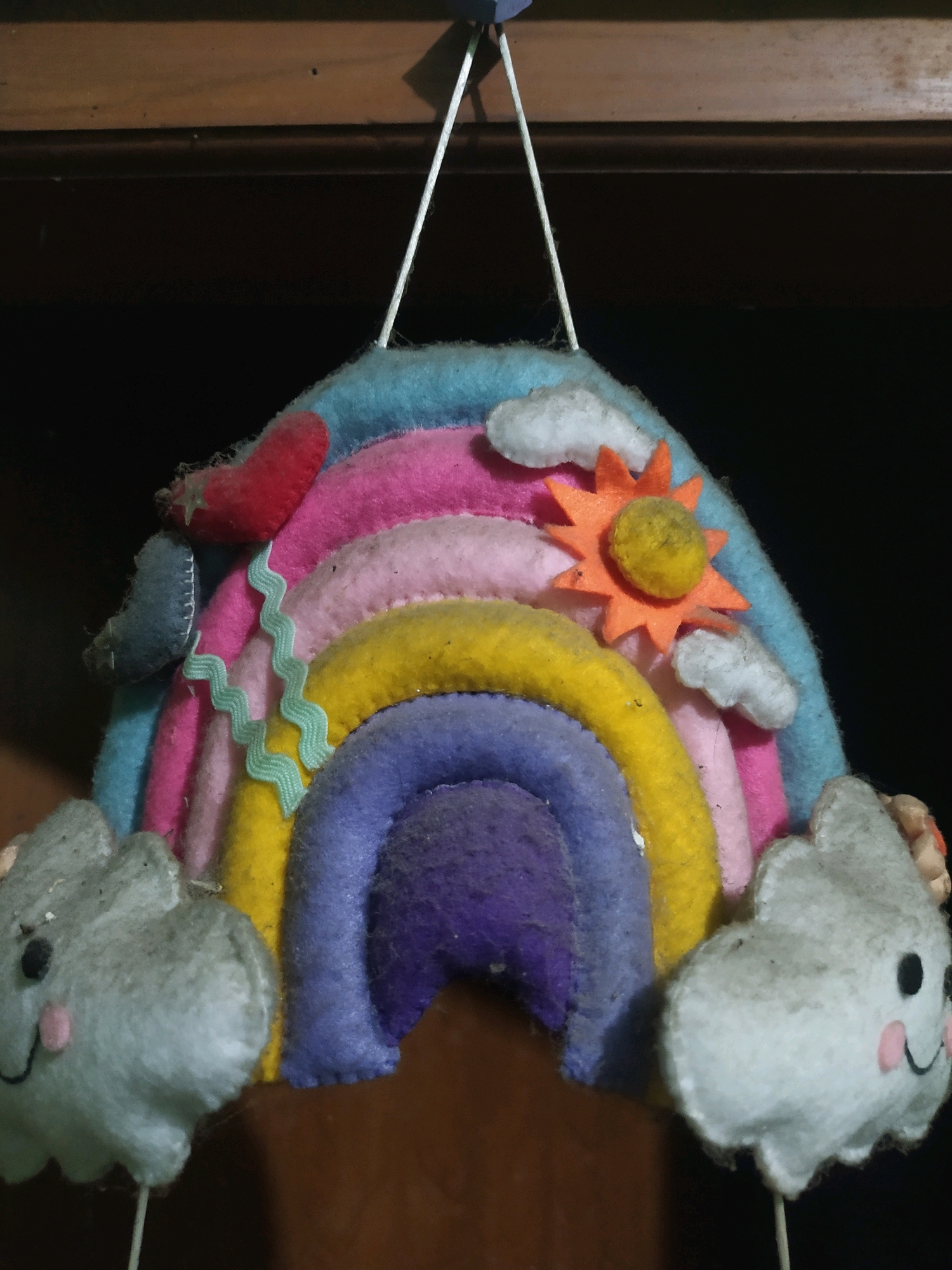 An indoor, eye-level, close-up shot focuses on a handmade felt rainbow decoration suspended from a dark wooden surface. The rainbow is crafted with multiple colored arcs, including blue, pink, yellow, and purple. Adorning the rainbow are small felt embellishments: a red heart, several light grey clouds with embroidered faces and pink cheeks, and an orange sun with a yellow center. A thin, light blue, zigzag ribbon is also attached to the rainbow. The background is dark, with the top of the frame showing a wooden plank. The overall impression is of a whimsical, handcrafted item. The image does not provide any information to determine a specific location within Sroyo, Indonesia.