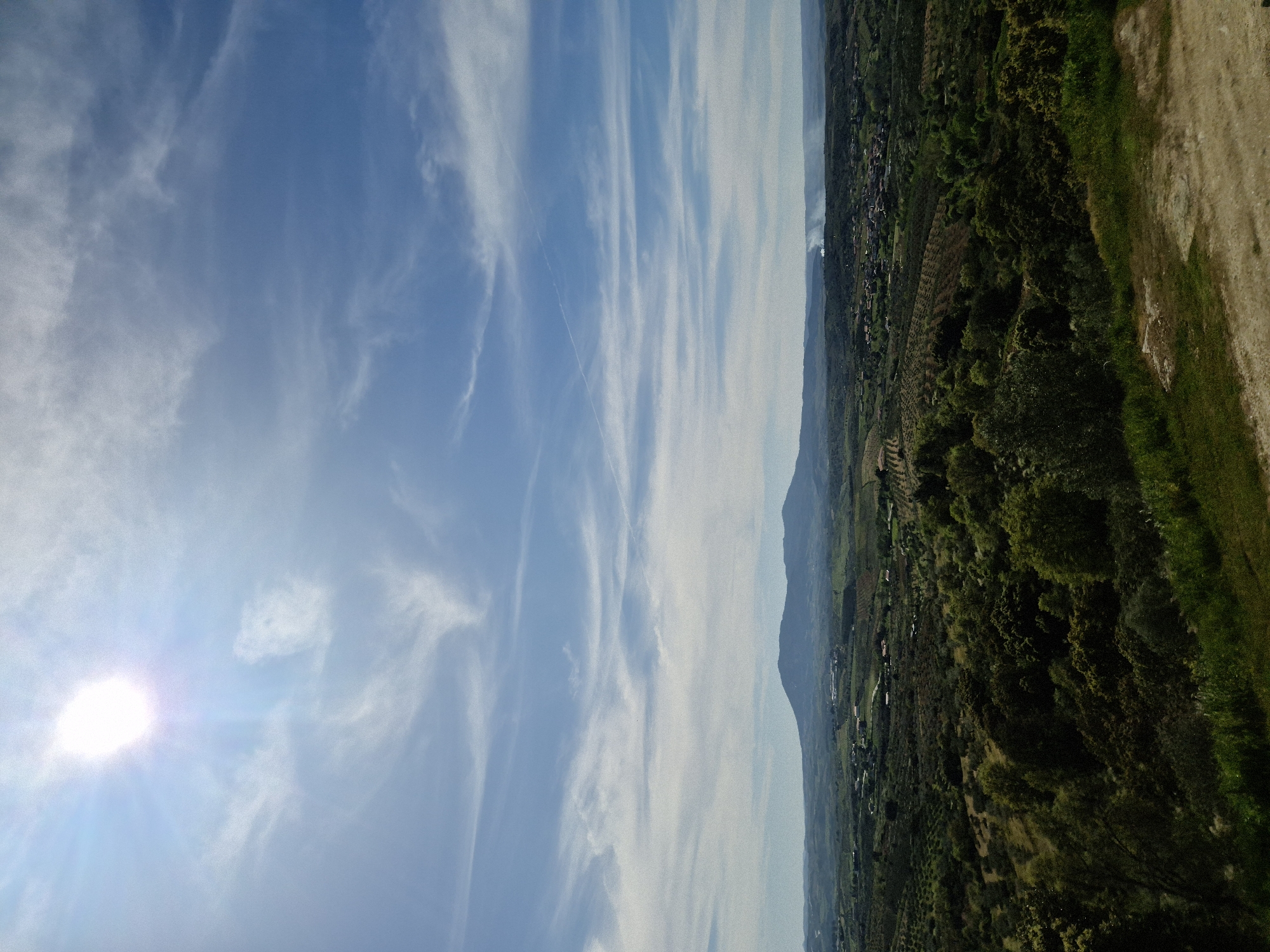 The image shows a landscape in Mirandela, Portugal, taken on a sunny day. The sky is blue with streaks of white clouds. The sun is shining brightly in the lower left corner. On the right side of the image, there's a view of land covered in green trees and vegetation, extending along a hillside. The terrain transitions from forest to a dirt path. A mountain range is visible in the distance separating the green landscape from the sky.
