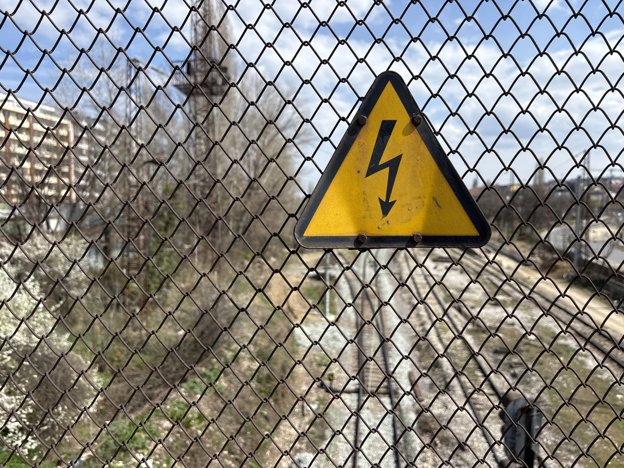 The scene depicts an urban railway corridor in Sofia, Bulgaria, viewed through a weathered, rust-brown chain-link fence. A faded yellow triangular warning sign, featuring a black border and a downward-pointing black lightning bolt symbol, is affixed to the fence slightly right of the frame's center with four visible rusty fasteners. Through the fence, under a partly cloudy blue sky, a blurred background reveals multiple parallel railway tracks with gravel ballast extending into the distance in the lower-right. On the left, a light-colored multi-story building with windows is partially visible. The midground contains numerous bare deciduous trees and shrubs. A tall, dark, skeletal structure, possibly a gantry crane or transmission tower, is discernible in the upper-mid background. A small, dark, indistinct figure or object is partially visible near the tracks in the lower-right. No specific actions or interactions are clearly discernible. The environment is characterized by industrial and urban infrastructure, separated by a security fence.