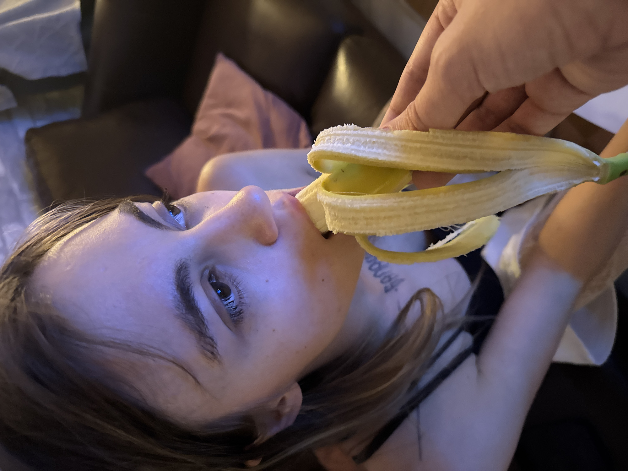 A close-up, low-light photograph captures a young woman indoors, appearing to be in Moscow, Russia, as she is being fed a peeled banana. The woman's face is partially visible, turned to the side, with her mouth open as she receives the banana. Her eyes are looking away from the camera, and her expression is neutral. She has dark hair, and a tattoo is visible on her neck. Her attire is a dark-colored tank top.
A hand, presumably belonging to another individual, is holding the banana and bringing it towards the woman's mouth. The banana is peeled, and the peel is partially unfurled, revealing the light yellow fruit. The background is dimly lit, suggesting an indoor environment, possibly a living room, with a dark-colored sofa and a pink cushion visible. The overall lighting is dim and casts a blueish hue on the scene. The setting does not provide any specific identifiers that would pinpoint the exact location within Moscow, Russia.