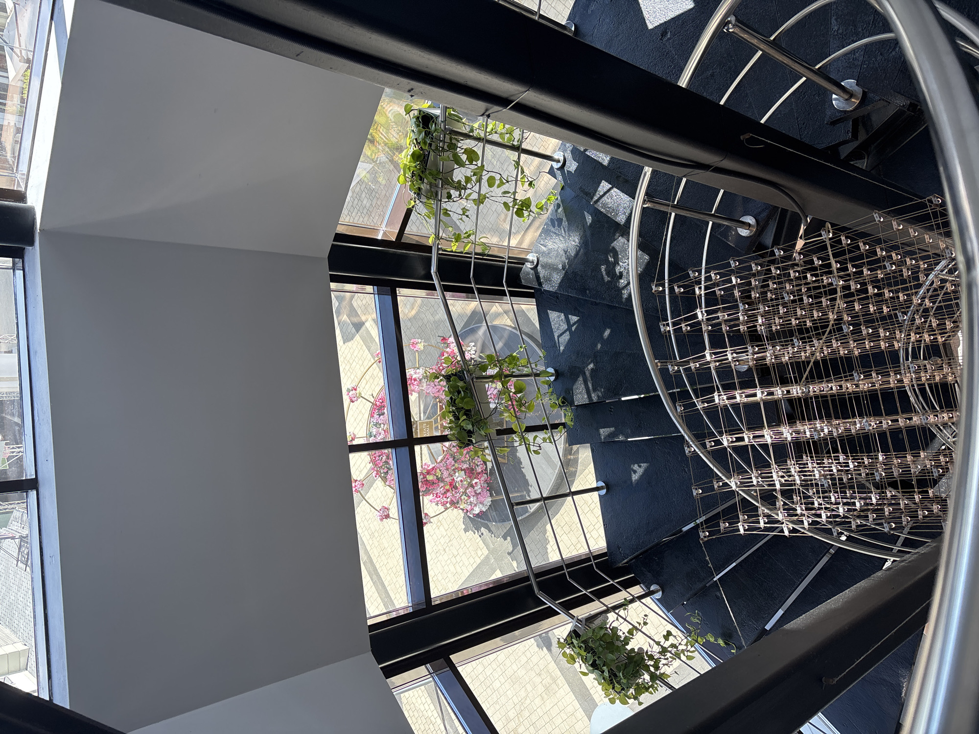 This is a high-angle, exterior shot looking down into a modern, multi-story building with a prominent spiral staircase in Monolit, Russia. The staircase has dark treads and is bordered by a sleek, stainless-steel railing. Along the railing, several potted plants with trailing green vines add a touch of nature to the metallic structure. Through large windows that form the exterior walls, the scene outside is visible, showing a paved courtyard with a decorative circular floral arrangement. Sunlight streams in, casting geometric shadows on the staircase and the floor below. A central, dark, structural pillar supports the building's upper levels. The overall impression is one of contemporary architecture blending indoor and outdoor elements.
