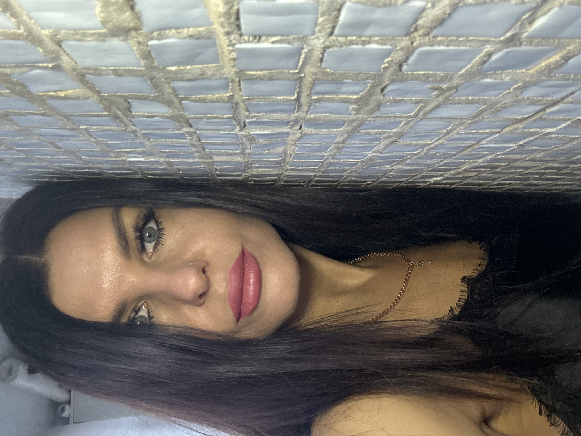 A woman with long dark hair, fair skin, and light eyes is positioned against a wall tiled with small, light-colored square tiles with dark grout. She is wearing a dark top with lace trim and a delicate gold necklace. Her lips are painted with a pinkish-red lipstick. The location is possibly in Chaikovsky, Russia.