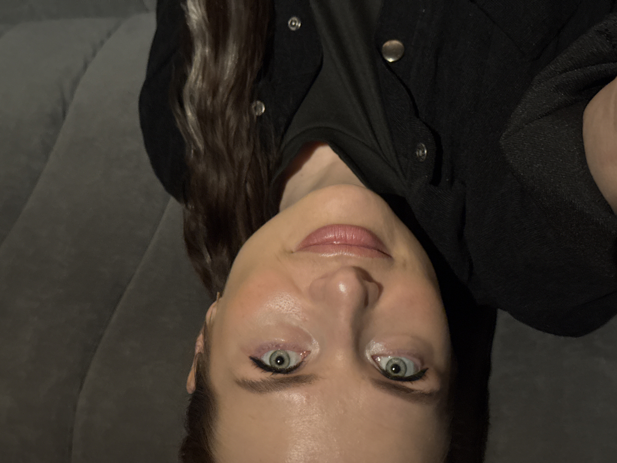 A single young adult female is depicted in a close-up portrait, positioned in a reclining or supine posture with her head tilted to the viewer's left. She gazes directly at the camera with a subtle, closed-lip smile. Her eyes are light blue-green, featuring black winged eyeliner and traces of pink eyeshadow. Her eyebrows are dark and well-defined, complementing her fair complexion. Her lips are full, glossy, and a muted pink shade. Dark, wavy hair with lighter brown or auburn highlights flows around her head. She is dressed in dark-colored clothing, including what appears to be a black crew-neck shirt beneath a dark jacket or overshirt detailed with multiple silver-toned metal snap buttons visible on the front placket and pocket flap. The background consists of a soft, textured gray fabric, likely an upholstered surface such as a sofa or bed, exhibiting gentle folds and contours. The lighting is dim and artificial, illuminating the subject's face from the front. The environment is indoors, but no specific geographic identifiers for the city of Chaikovsky, Russia, or any other location are discernible within the image.