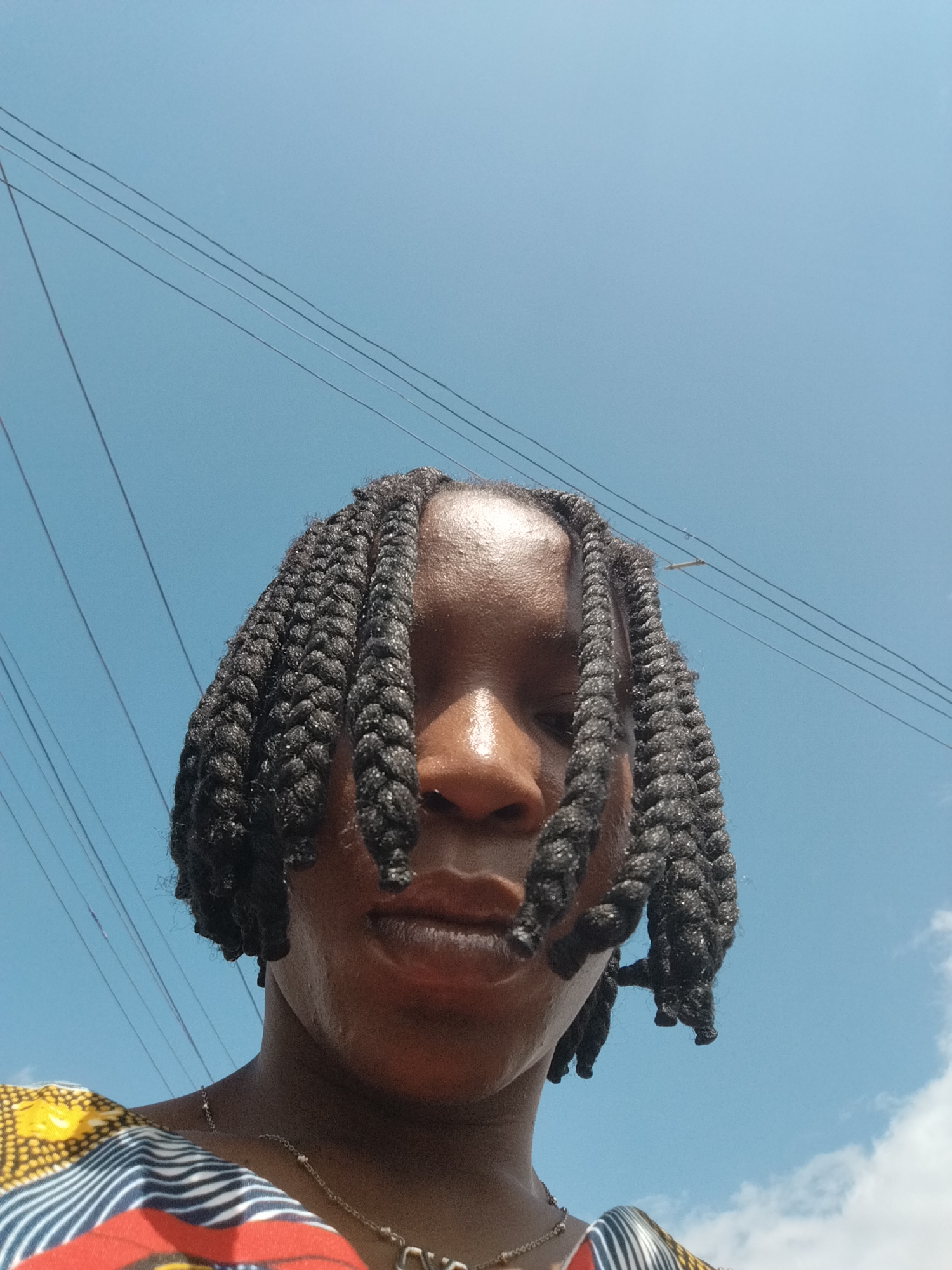 The image is a close-up, low-angle shot of a person with braided hair against a bright blue sky with scattered white clouds. The person's face is partially obscured by their braids, with only their forehead, nose, mouth, and chin visible. Their skin appears to be glistening, suggesting it is either sweaty or oily. They are wearing a colorful garment with a patterned design, and a thin silver necklace is visible around their neck. In the background, several power lines are stretched across the sky. The overall lighting suggests it is daytime, likely outdoors. The image does not provide specific visual cues to pinpoint the exact location within Isiehor, Nigeria, other than it is an outdoor setting with infrastructure for electricity.