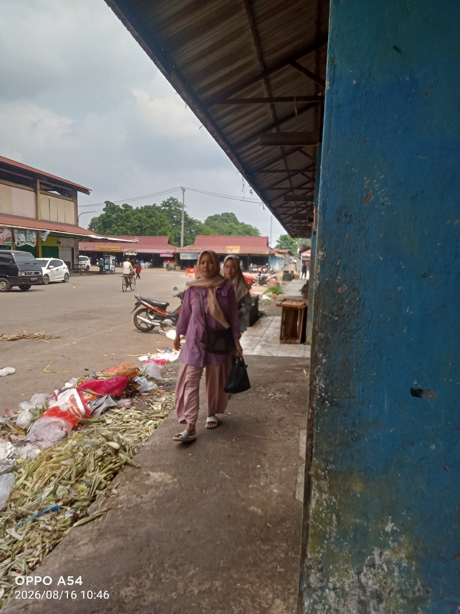 A full shot captures a busy street scene in Jambi, Indonesia, on an overcast day. In the foreground, two women walk along a sidewalk lined with a large pile of garbage and discarded plant matter. The woman in the foreground wears a purple headscarf and a loose-fitting purple top and light pink pants. She carries a black handbag and walks with her head down. Behind her, another woman, also wearing a headscarf, walks with her head slightly turned. A blue wall with peeling paint occupies the right side of the frame. In the background, several market stalls with red roofs are visible, along with parked motorcycles and cars. A man rides a bicycle across the street. The street appears to be a public market area, characterized by its open-air stalls and the presence of numerous people and vehicles.
