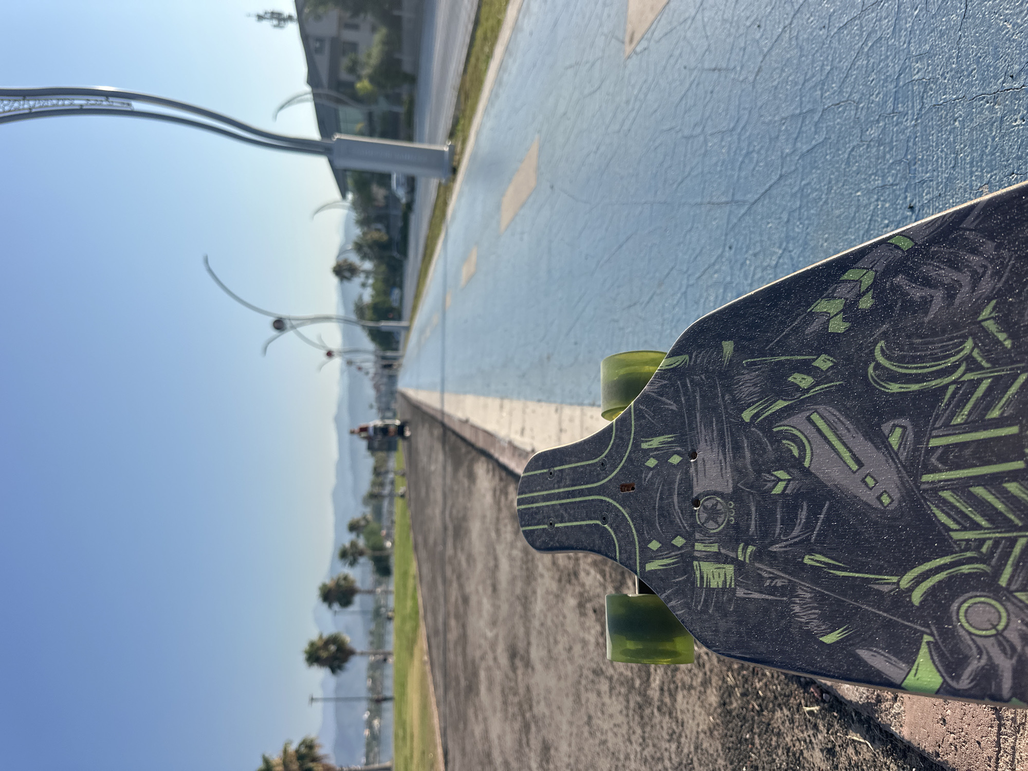 A first-person perspective shot shows a longboard with a black and green graphic design resting on a paved path adjacent to a blue bike lane. The scene is set in Fethiye, Turkey, with palm trees, a body of water, and mountains visible in the background under a clear blue sky. In the distance, a person is riding a scooter or bicycle on the paved path. The overall atmosphere suggests a recreational outdoor setting in a coastal city.