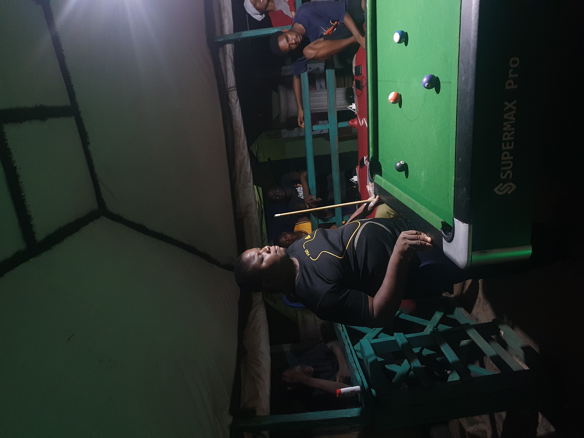The image shows a group of young men in an outdoor setting, likely in Lokoja, Nigeria, at night. The primary focus is a billiards table, with several balls scattered across its green surface. One man is actively playing, holding a cue stick and positioned to strike a ball. He is wearing a black t-shirt with yellow detailing. Other individuals are visible around the table, some watching the game and others engaged in conversation or relaxation. The scene appears to be dimly lit, with artificial light sources creating shadows and highlighting the playing area. There are some structures in the background, including what looks like a corrugated metal roof and some wooden beams, suggesting a casual or informal establishment. The overall atmosphere seems relaxed and social, characteristic of a gathering of friends. The text "SUPERMAX Pro" is visible on the side of the billiards table. The time of day is clearly night, and there are no indicators of weather conditions.