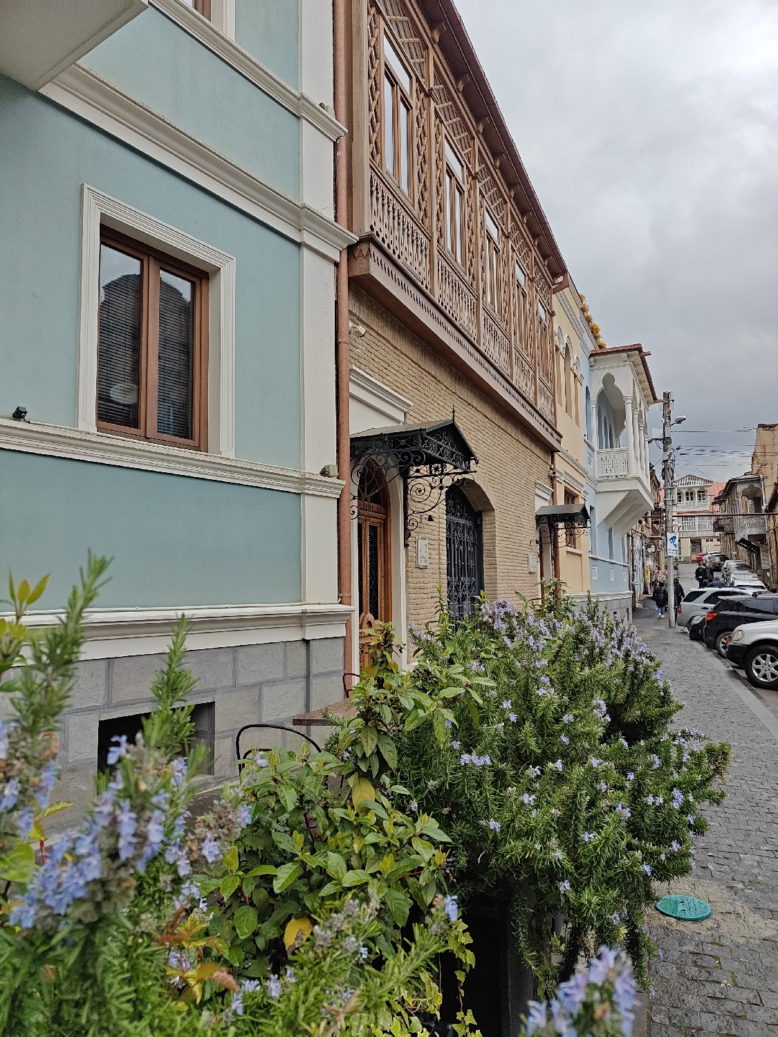This media file captures a charming street scene in Tbilisi, Georgia, featuring traditional architecture and lush greenery under an overcast sky.
In the foreground on the left, vibrant green bushes with small, delicate purple-blue flowers partially obscure the view. Behind these plants stands a building with a light blue-green facade, accented by white decorative trim around a prominent wooden-framed window, and a grey stone-block base.
Adjacent to it, a second building displays a light brown brick ground floor topped by an ornate, intricately carved wooden balcony or gallery on the second story. This balcony features multiple windows and detailed railings. Below it, a dark, decorative wrought-iron awning with elaborate scrollwork shelters an entrance. Further along the street, more colorful buildings, some with traditional white carved balconies, line the right side.
The street itself is paved with cobblestones. Several cars are parked bumper-to-bumper along the right side, stretching into the distance where the street gently curves. Utility poles and overhead wires are visible above the parked vehicles. A green manhole cover is visible on the cobblestone street. In the distance, a few indistinct figures can be seen walking on the sidewalk. The sky is uniformly cloudy, suggesting an overcast day. No discernible text is visible on any signs or buildings. The image conveys the historic charm and lively atmosphere of a European city street.