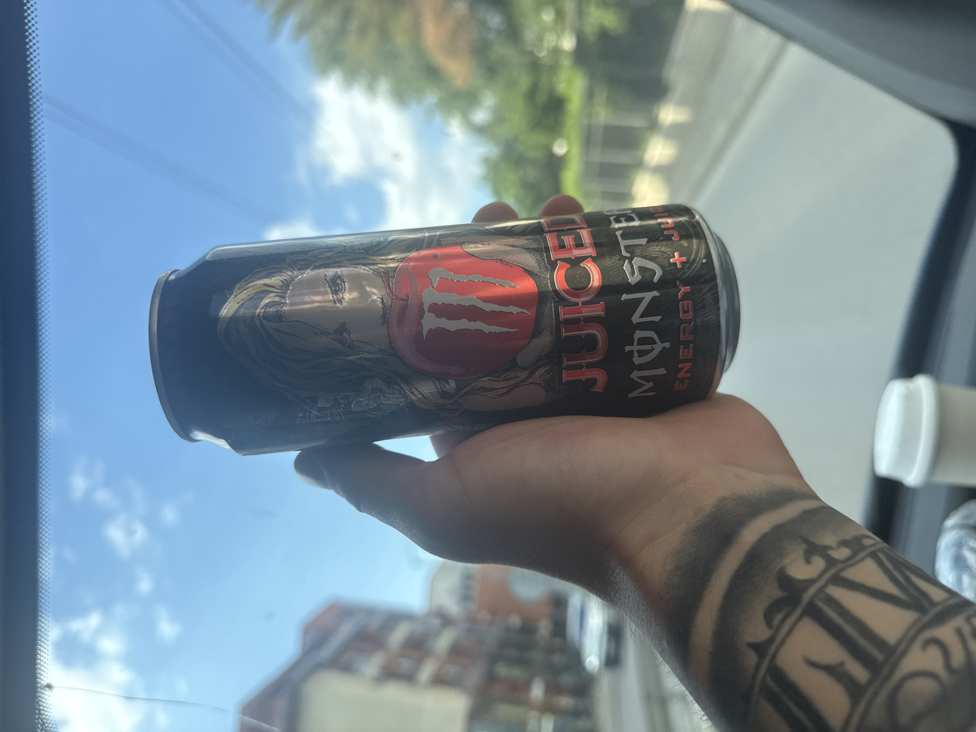A person with a tattooed forearm is holding a can of "Juiced Monster" energy drink. The background shows a city scene with buildings and a blue sky with clouds. The location is likely Sofia, Bulgaria, based on the architecture and appearance of the buildings. The photograph was taken inside a vehicle, as evident by the car window frame.