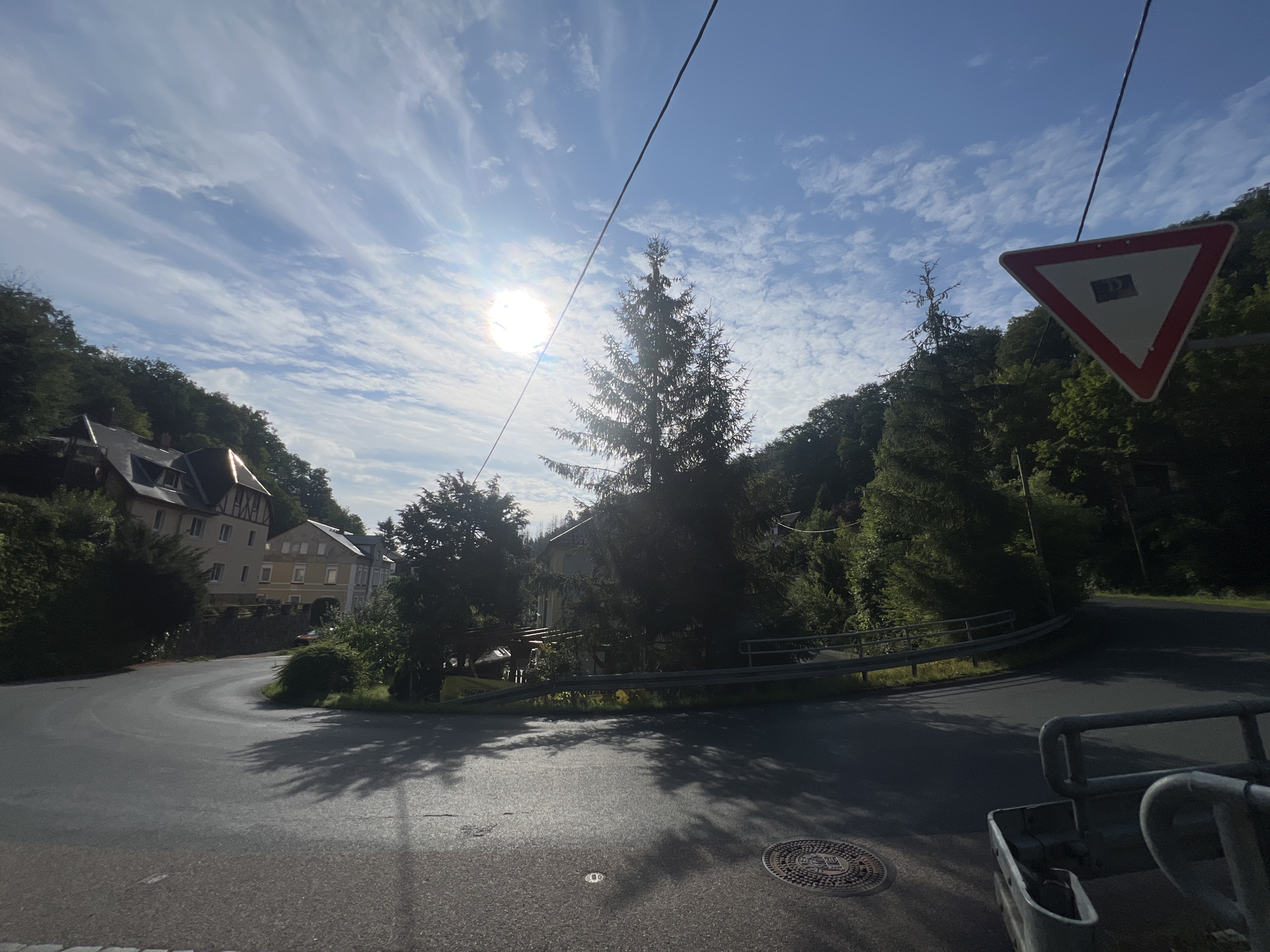 The image captures a view of a road in the city of Freital, Germany, on a sunny day with scattered clouds. The road curves to the left, with trees and houses lining the hillside. A prominent triangular yield sign with a red border and a white interior displaying a black symbol is visible in the upper right portion of the frame. In the distance, several buildings with traditional European architecture, featuring dark roofs and light-colored facades, are nestled among the trees. A tall evergreen tree stands near the center of the image, silhouetted against the bright sun. The sun is high in the sky, creating lens flare and casting shadows on the road. The foreground shows the asphalt surface of the road, with a manhole cover and a metallic railing on the right side. The environment appears to be a semi-rural or suburban area with a blend of residential structures and natural landscape.