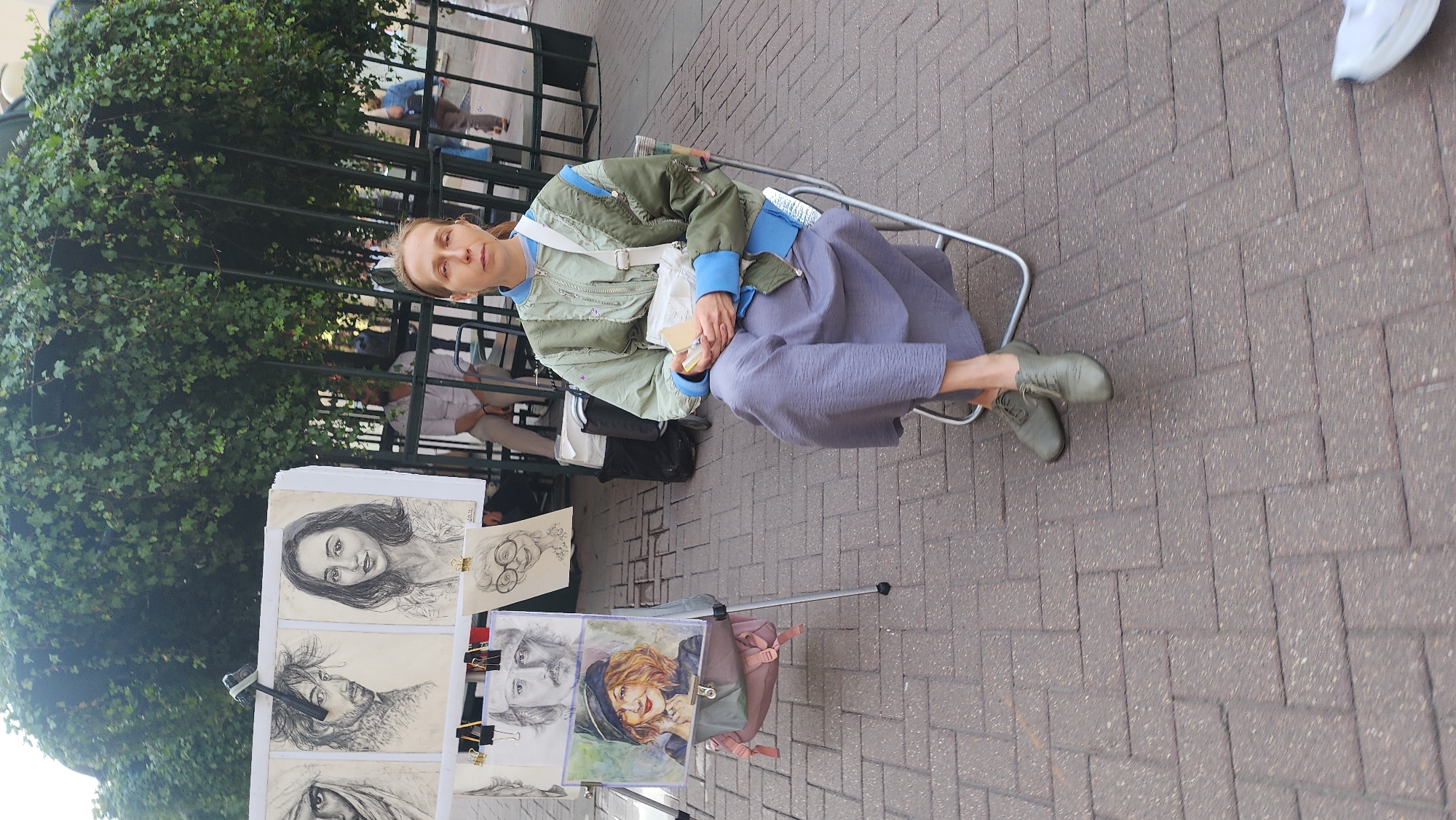 The media file shows a woman sitting in a chair outdoors, in front of a display of artwork. She is wearing a green jacket with blue accents and a long, grey skirt. Her feet are resting on the ground, and she is holding a small object in her hands. The artwork displayed in front of her consists of several portraits, seemingly sketches and paintings.
The scene appears to be a street or public walkway, with a brick-paved ground. A metal fence with green foliage behind it is visible to the left of the woman. In the background, other people can be seen, some standing and others sitting. The lighting suggests it is daytime, with bright sunlight casting shadows.
The woman's expression is neutral, and the overall atmosphere seems relaxed, suggesting a typical day in a public space where art is being displayed or sold. While specific location cues for Moscow are not immediately obvious in the image itself, the context provided indicates this setting. No visible text is discernible in the media file.
