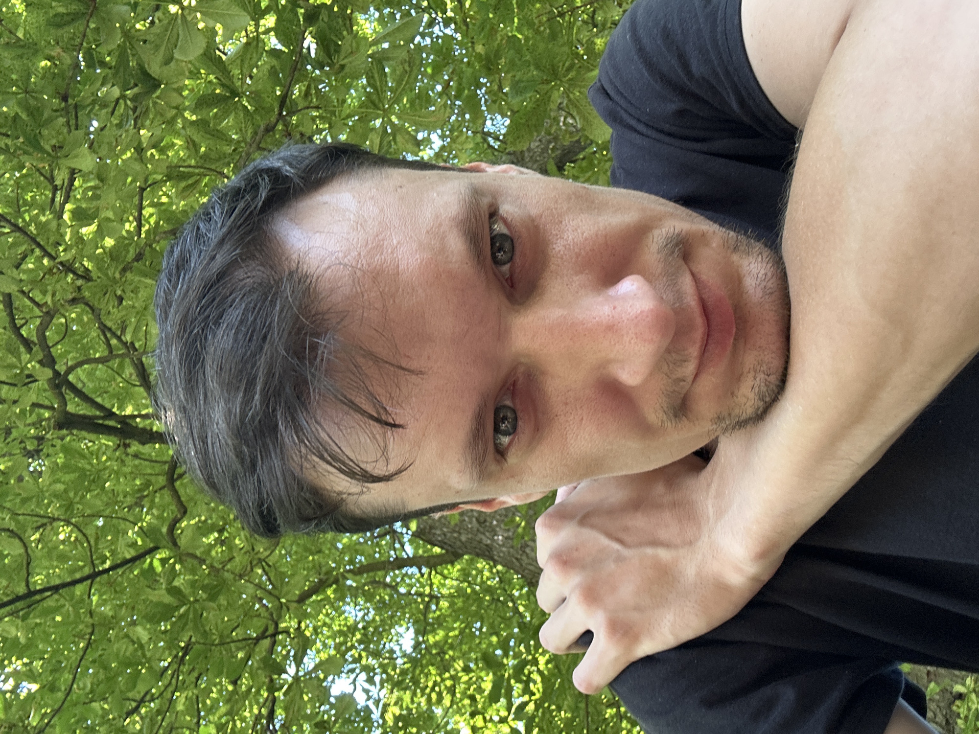 A close-up, selfie-style photograph shows a man in the foreground, with his face tilted slightly downwards and to the right. He has dark hair, light skin, and blue eyes. His left arm is bent, with his forearm resting on his chest and his hand grasping his upper left arm. He is wearing a black t-shirt. Behind him, out of focus, is lush green foliage, suggesting an outdoor setting with trees. The image is taken from a low angle, looking up at the man. No specific location in Makiv, Ukraine, can be identified from the visual information provided.