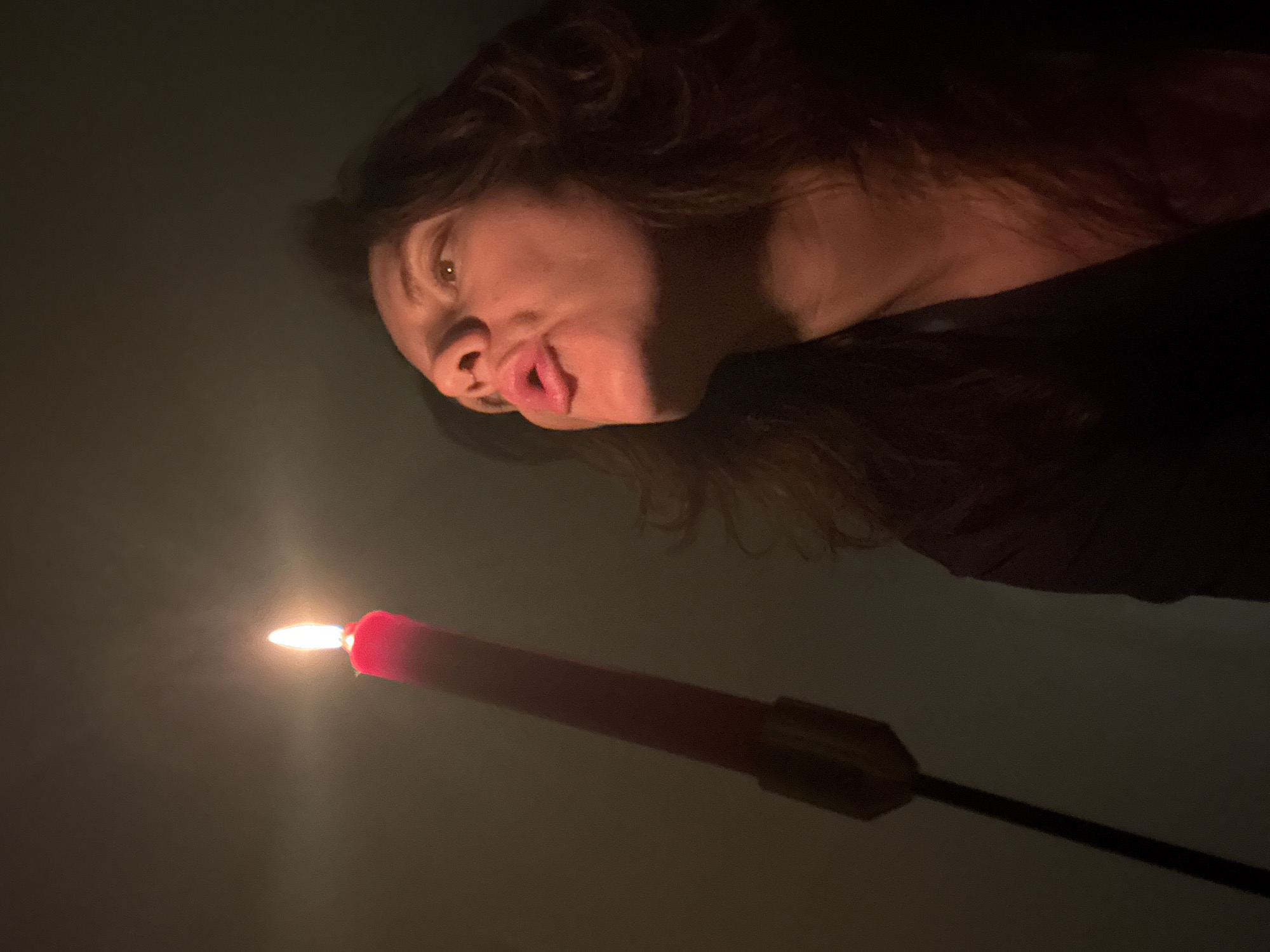 A woman with dark, curly hair is positioned at the top of the frame, her face illuminated by the light of a red candle with a bright flame. Her lips are pursed as if she is about to blow out the candle. The candle is held in a metal holder attached to a black rod, angled upwards towards the woman. The background is dark and indistinct. There is no evidence to confirm the location is Boreti, Montenegro.