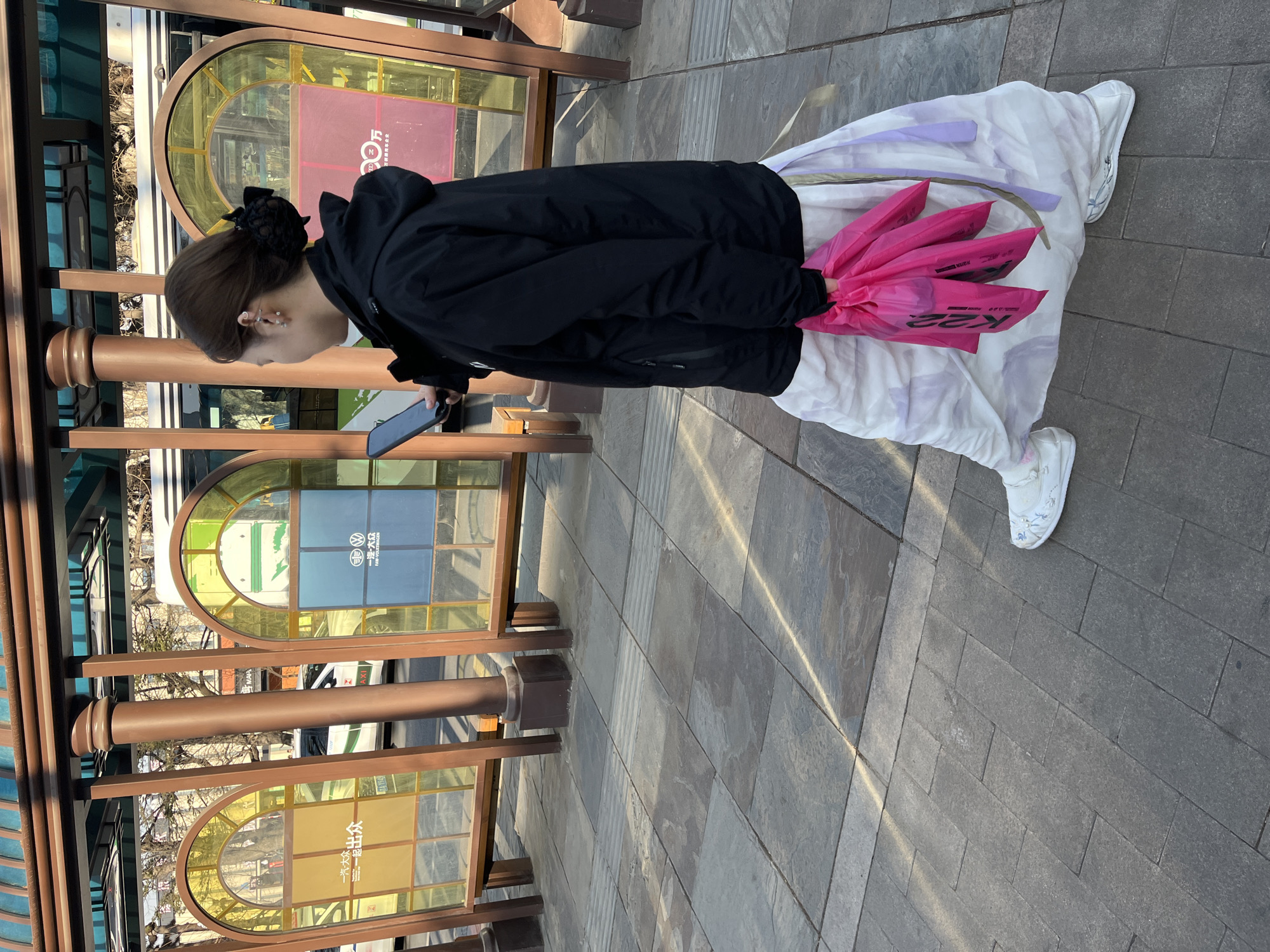 A woman stands on a paved walkway adjacent to a bus shelter in Changchun City, China. She is wearing a black jacket, a long white skirt with a light purple pattern, and white sneakers. Her dark hair is styled with a black hair accessory. She holds a cell phone in her left hand and a bundle of four bright pink plastic shopping bags in her right hand. The bus shelter has a distinctive design with bronze metal framing and multiple sections of arched, colored glass panels. Sunlight casts shadows across the walkway.