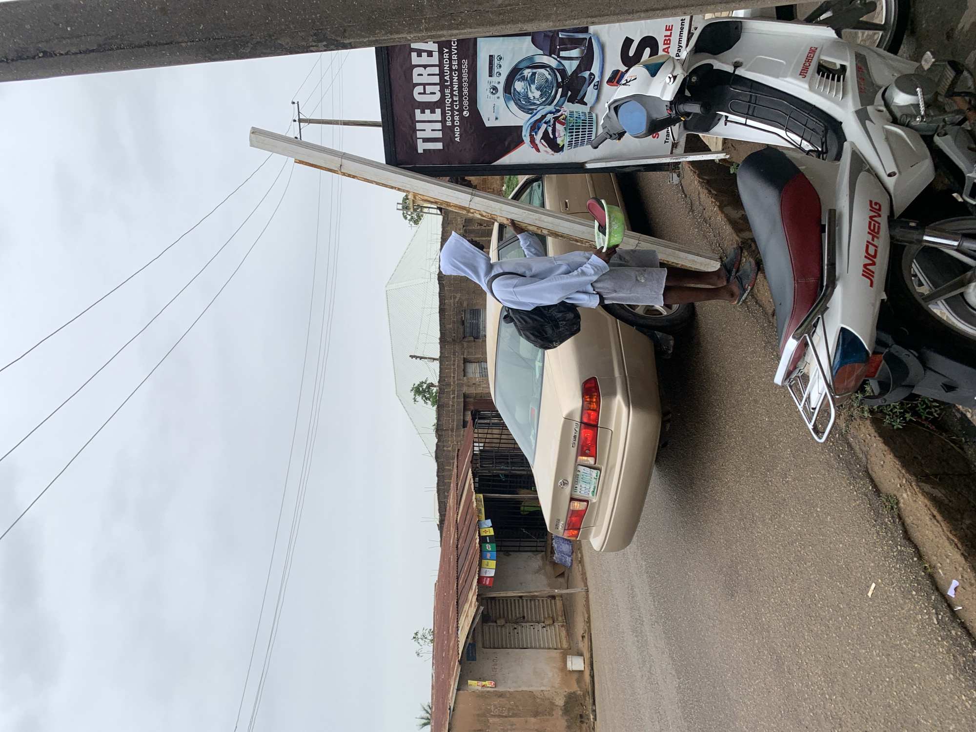 A daytime street scene in what appears to be a commercial/residential area. A man in a hooded sweatshirt, shorts, and sandals stands near a beige car, holding a green object. A white motorcycle is parked beside the car. A sign for "The Great Boutique, Laundry, and Dry Cleaning Service" is visible, along with a phone number. A utility pole and overhead wires are present. Across the street is a weathered building with a corrugated roof and closed storefronts. The car's license plate reads "GGE-40340". Based on the phone number on the sign, this is likely in Nigeria. The brand name "JINCHENG" appears on the motorcycle. The location is plausibly Jos, Nigeria.