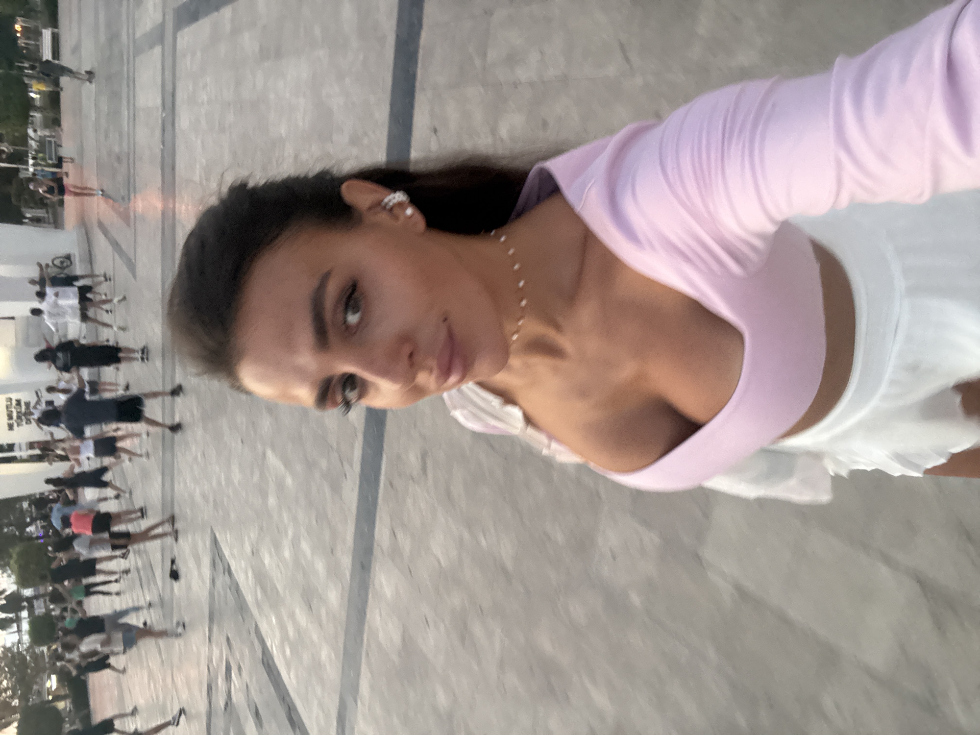 In Fethiye, Turkey, a young woman takes a selfie in a public square. She is wearing a pink long-sleeved shirt and a white ruffled skirt. In the background, a group of people are engaged in an outdoor exercise class. The square is paved with large stone tiles, and there are some trees and buildings visible in the distance. The sun is setting, casting a warm glow over the scene.