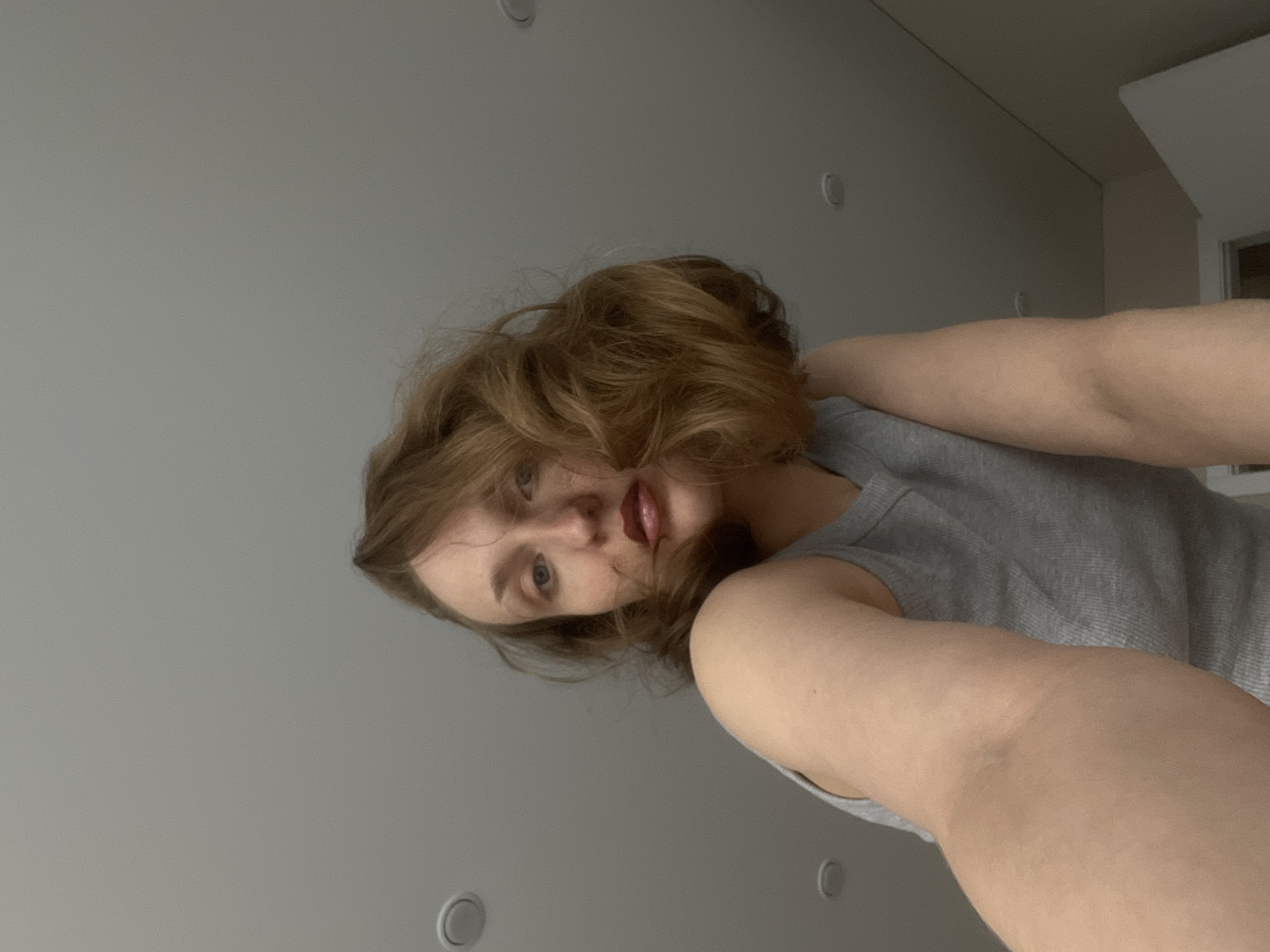 A woman with shoulder-length, light brown hair and fair skin is wearing a gray tank top and is looking directly at the camera. Her lips are painted a dark red. The background is a light gray wall with recessed ceiling lights. The location in Подшивалово, Russia cannot be determined based on the image alone.