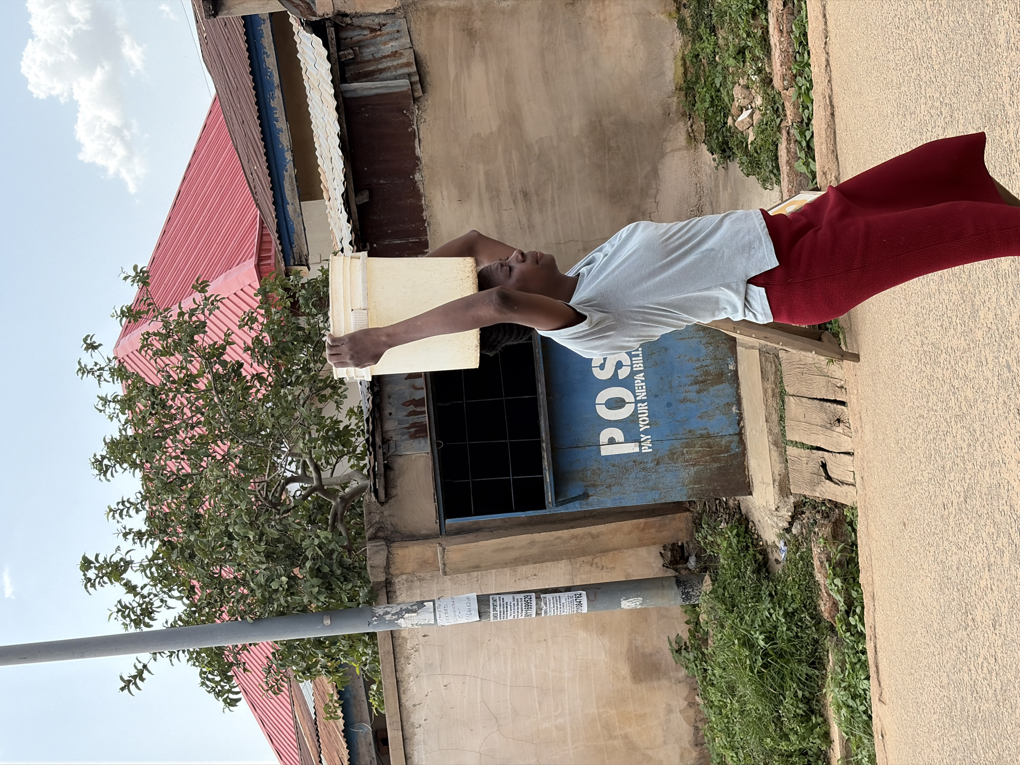 A young person with brown skin, dressed in a light-colored short-sleeved shirt and maroon pants, lies horizontally with their arms extended above their head. Their head rests on a stack of light-colored buckets positioned on top of a blue metal mailbox or distribution box labeled "POS" with the phrase "PAY YOUR NEPA BILLS" below it. The mailbox is attached to a weathered building with a dark-framed window. To the left, there is a pole with multiple small printed notices affixed to it. A leafy tree is also visible on the left side of the frame, partially obscuring the red corrugated roof of a nearby building. The background includes parts of buildings, overgrown vegetation, and a paved surface. The scene appears to be an outdoor urban setting in Jos, Nigeria.
