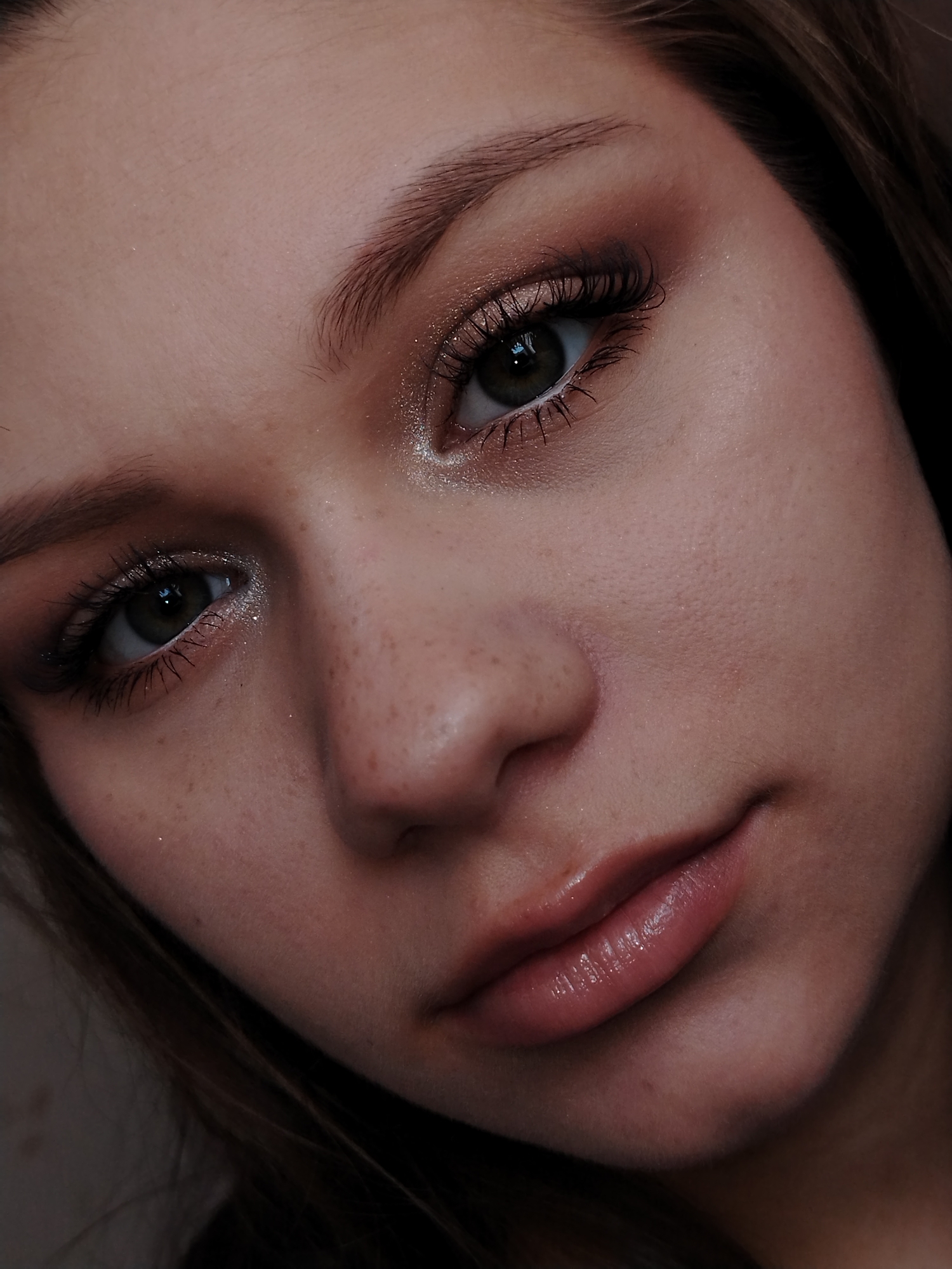 A close-up portrait shot features a young person, likely female, from the forehead to the chin, with a portion of dark brown hair visible on the right side. The person is looking directly at the camera with a neutral to soft expression, a slight hint of a smile playing on the lips.
The eyes are a prominent feature, appearing dark green or hazel, framed by long, dark eyelashes, suggesting mascara or false lashes. The eyelids are adorned with makeup, including a shimmery, light-colored eyeshadow on the inner corners and lower lash line, complemented by a darker, warm-toned (brown/gold) eyeshadow blended across the lids and outer corners. The eyebrows are well-defined. Visible freckles dot the bridge of the nose and cheeks, adding to the natural skin texture, where pores are also subtly visible. The skin also shows a subtle shimmer, possibly from a highlighter, on the cheekbones and forehead. The lips are full and appear to be coated in a glossy, natural pink or nude-toned product.
The lighting is soft and diffused, highlighting the facial features and the shimmer of the makeup. The background is indistinct and dark, keeping the focus entirely on the subject. There is no visible text in the image. The scene is likely an indoor setting, given the controlled lighting and close-up nature of the portrait.