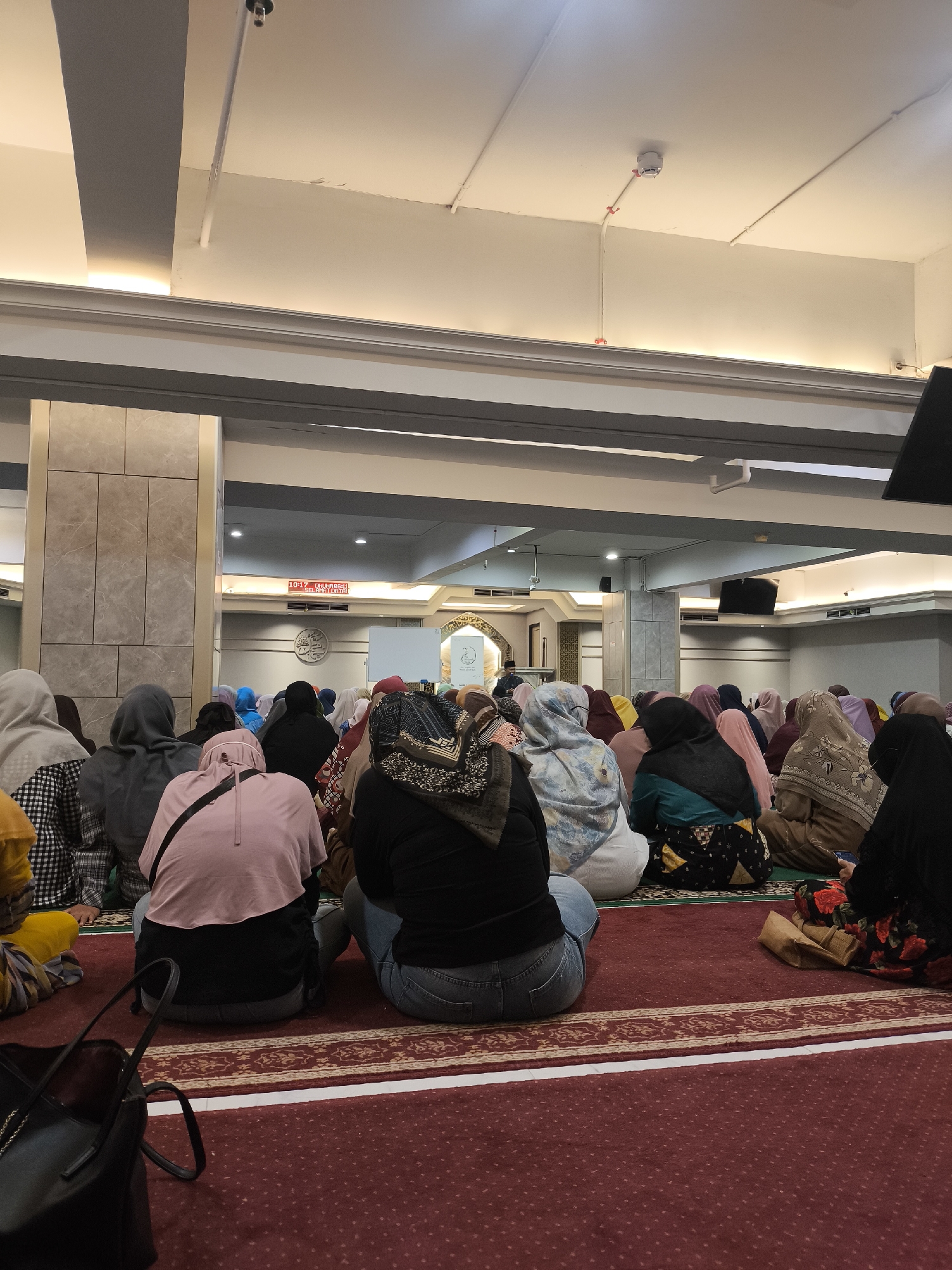In Grogol, Indonesia, an indoor scene depicts a group of people, predominantly women, sitting on a red carpet in what appears to be a place of worship. They are all facing away from the camera and towards a focal point in the distance, suggesting a prayer or religious gathering. The individuals are wearing various headscarves and modest clothing. The environment is illuminated by artificial lighting, with visible architectural features such as columns, a ceiling with exposed conduits, and decorative wall panels. A digital display screen showing "10:17" is also visible on a wall in the background. The overall atmosphere is one of quiet contemplation and community.