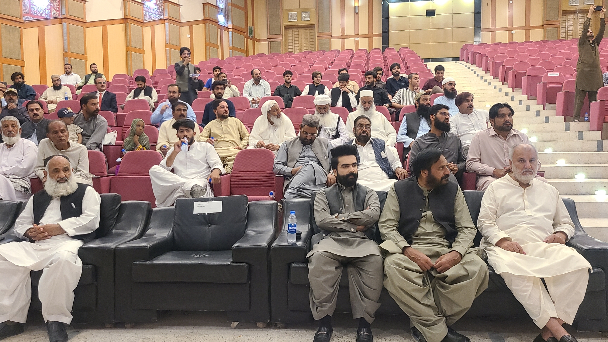 The image shows a seated audience in an auditorium, likely in Quetta City Tehsil, Pakistan. Most attendees are men dressed in traditional Pakistani clothing, including shalwar kameez and vests. They are seated in maroon-colored auditorium seats and on black sofas in the front row. A man stands on the stairs to the right, holding up a phone as if taking a picture or video. Another man in the middle rows is using a microphone. A bottle of water is visible on a black sofa between two people. The hall has wooden paneling on the walls and a stepped seating arrangement.