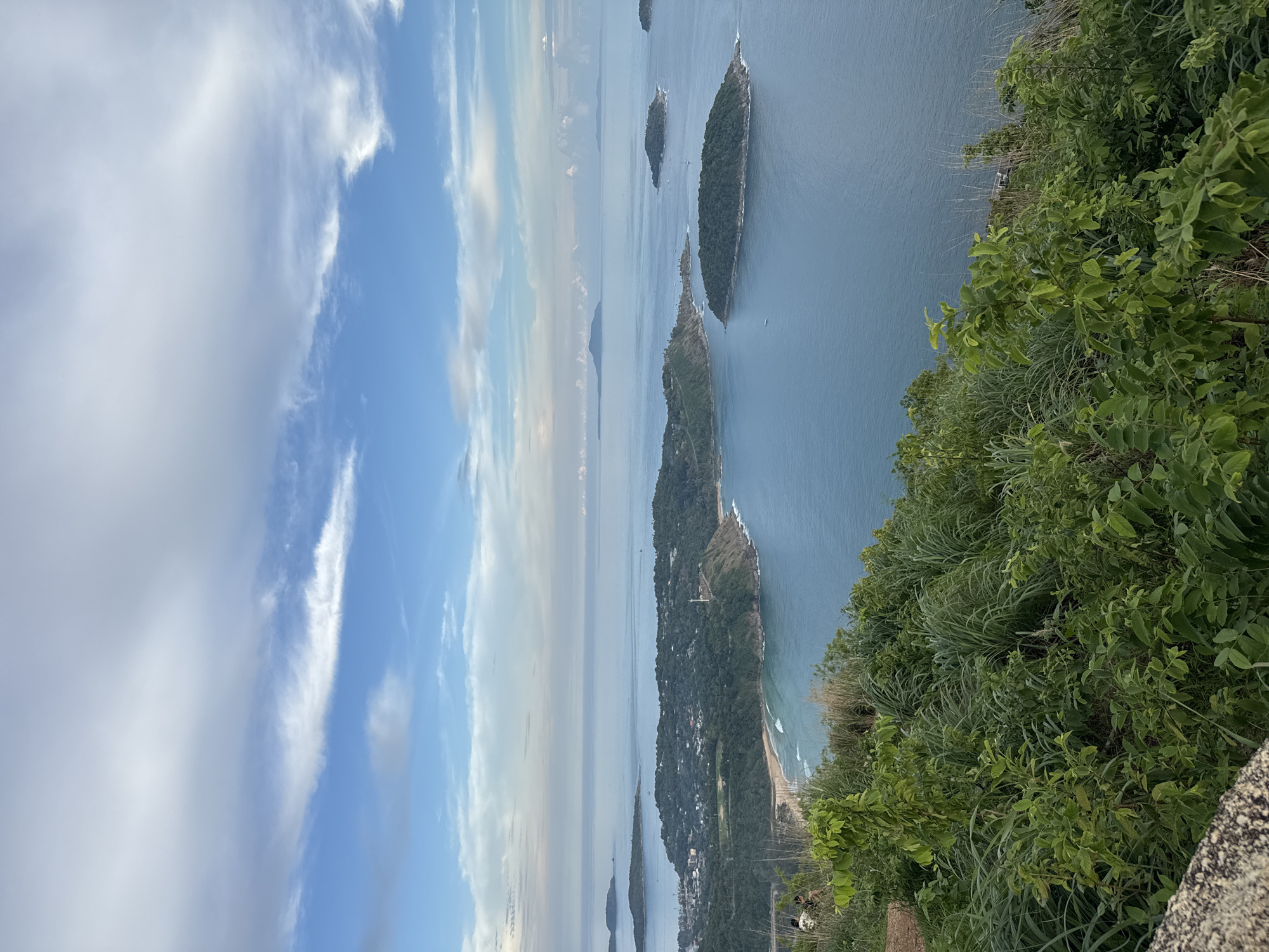 This is an aerial shot of a coastal landscape in Karon, Thailand, likely taken from a high vantage point. The scene is dominated by the vast blue ocean, dotted with several green islands of various sizes. In the foreground, lush green vegetation, including bushes and tall grasses, covers the hillside, with a stone ledge visible on the left.
The middle ground showcases the coastline of Karon, featuring a sandy beach with gentle waves lapping at its shore. A prominent lighthouse stands on a green, tree-covered headland that juts out into the sea. The land is densely forested, with some buildings visible near the coast. Two individuals are seated on the grassy slope in the lower left corner of the frame, facing away from the camera and towards the ocean view.
The background is characterized by the horizon where the sea meets the sky. The sky is a mix of blue and white clouds, with the clouds appearing lighter and more dispersed further away. The overall impression is a serene and picturesque view of a tropical coastline.