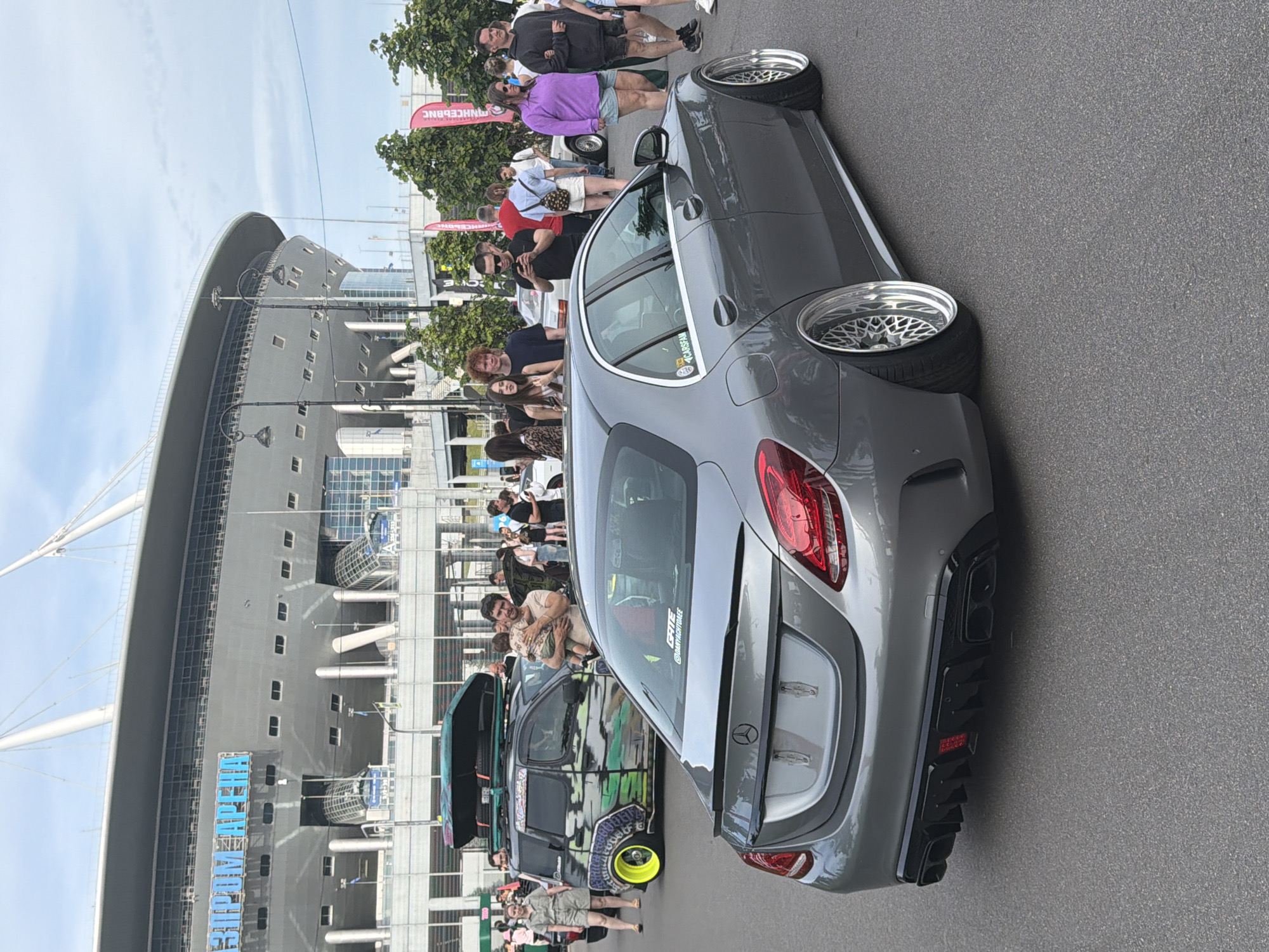 A grey Mercedes-Benz C-Class sedan is parked on asphalt in front of a stadium with the text "ГАЗПРОМ АРЕНА" visible on its facade. Several people are standing around the car and in the background, some are looking at their phones. A black roof box is attached to a black van with custom green and black graphics parked to the left of the Mercedes. The stadium has a modern architectural design with a curved roof supported by white struts. The sky is partly cloudy.