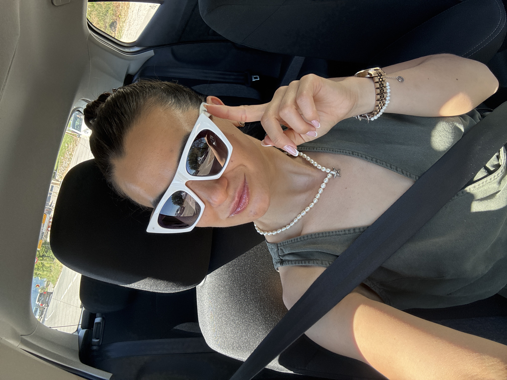A woman with her hair pulled back in a bun and wearing white cat-eye sunglasses, a pearl necklace, and a green sleeveless top is taking a selfie in the passenger seat of a car. She is wearing a seatbelt. Her left arm is visible, resting on her lap. On her right wrist, she wears a watch and a pearl bracelet with a small charm. The background shows the interior of a car, with a glimpse of a road and trees through the rear window. The location is Blizikuće, Montenegro.