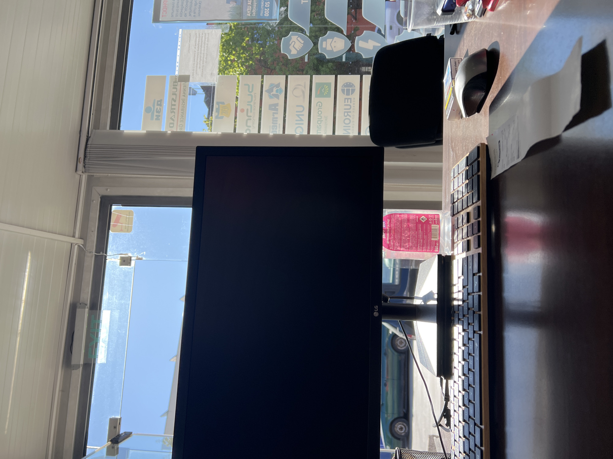 The image captures an indoor scene in Sofia, Bulgaria, as indicated by the prominent display of Bulgarian insurance company logos in the background window. Dominating the foreground is an LG computer monitor, a keyboard, and a computer mouse on a dark wooden desk. A black office chair is positioned in front of the desk. The window reveals a clear, sunny day with the sky visible above. A green vehicle is partially visible through the glass door. The blinds are partially closed. A bottle of disinfectant is also visible on the desk.