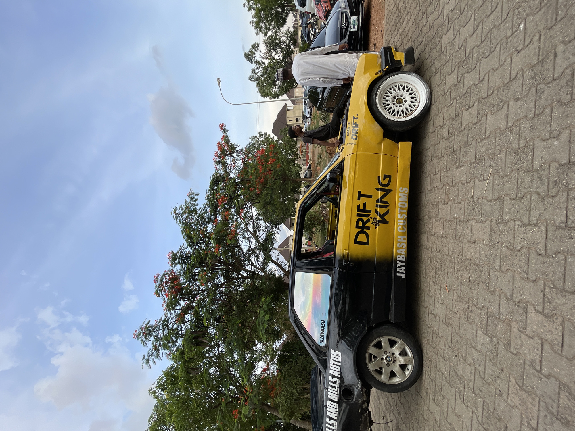 The image shows a black and yellow car with the words "DRIFT KING" and "JAYBASH CUSTOMS" on its side parked on a brick pavement. Two men are standing near the car, one wearing a white robe and hat, and the other in a darker outfit. Other cars are visible in the background. A tree with red flowers is near the car. The sky is blue with clouds. The location is likely in Abuja, Nigeria.