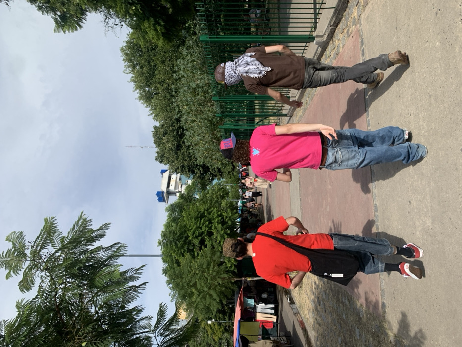 Three people are standing on a sidewalk next to a park. The first person wears a brown shirt, jeans, and a headscarf. The second wears a pink shirt, jeans, and a baseball cap. The third wears a red shirt, jeans, and a black bag slung over their shoulder. They are all facing the same direction. There are trees, a green fence, a building, and the sky in the background.