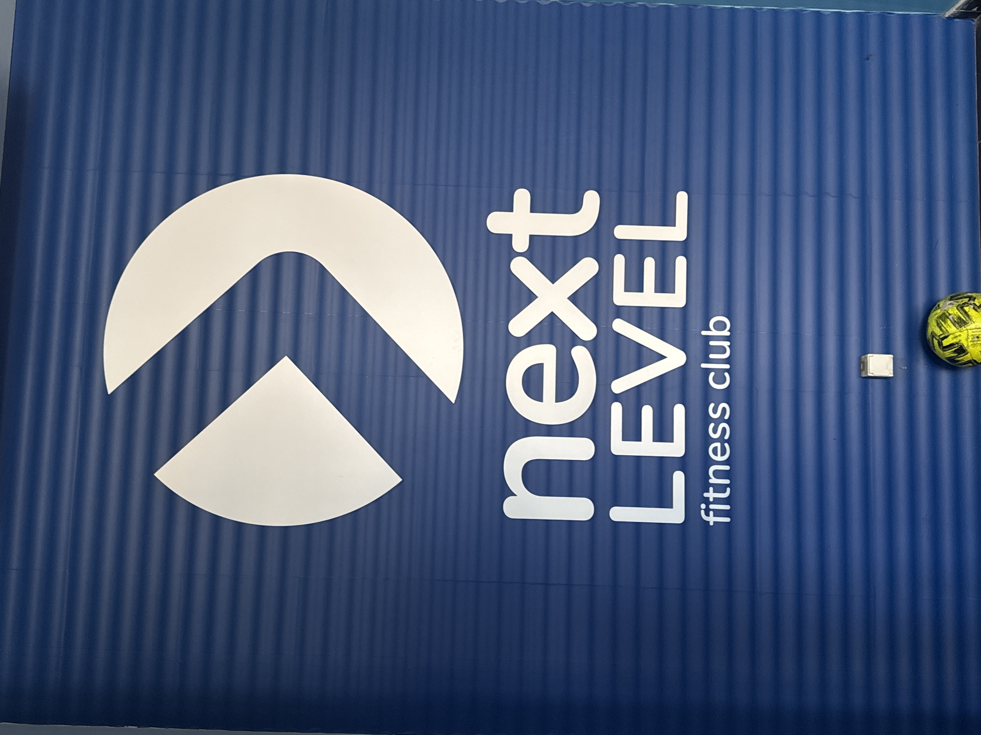 A blue, corrugated wall displays a white logo and text. The logo is an abstract design featuring a large curved shape resembling an open arrow or a curved arrow, with a smaller triangle inside its opening. Below the logo, the words "next LEVEL" are written in large, white, sans-serif capital letters, with "next" above "LEVEL". Underneath this, in smaller white text, are the words "fitness club". At the bottom of the image, a small square white electrical outlet or light switch is visible on the wall, and a yellow and black patterned ball rests on the floor in front of the wall. The scene is indoors, likely in a gym or fitness center, with the branding "next LEVEL fitness club" prominently displayed. The location is identified as Sofia, Bulgaria.