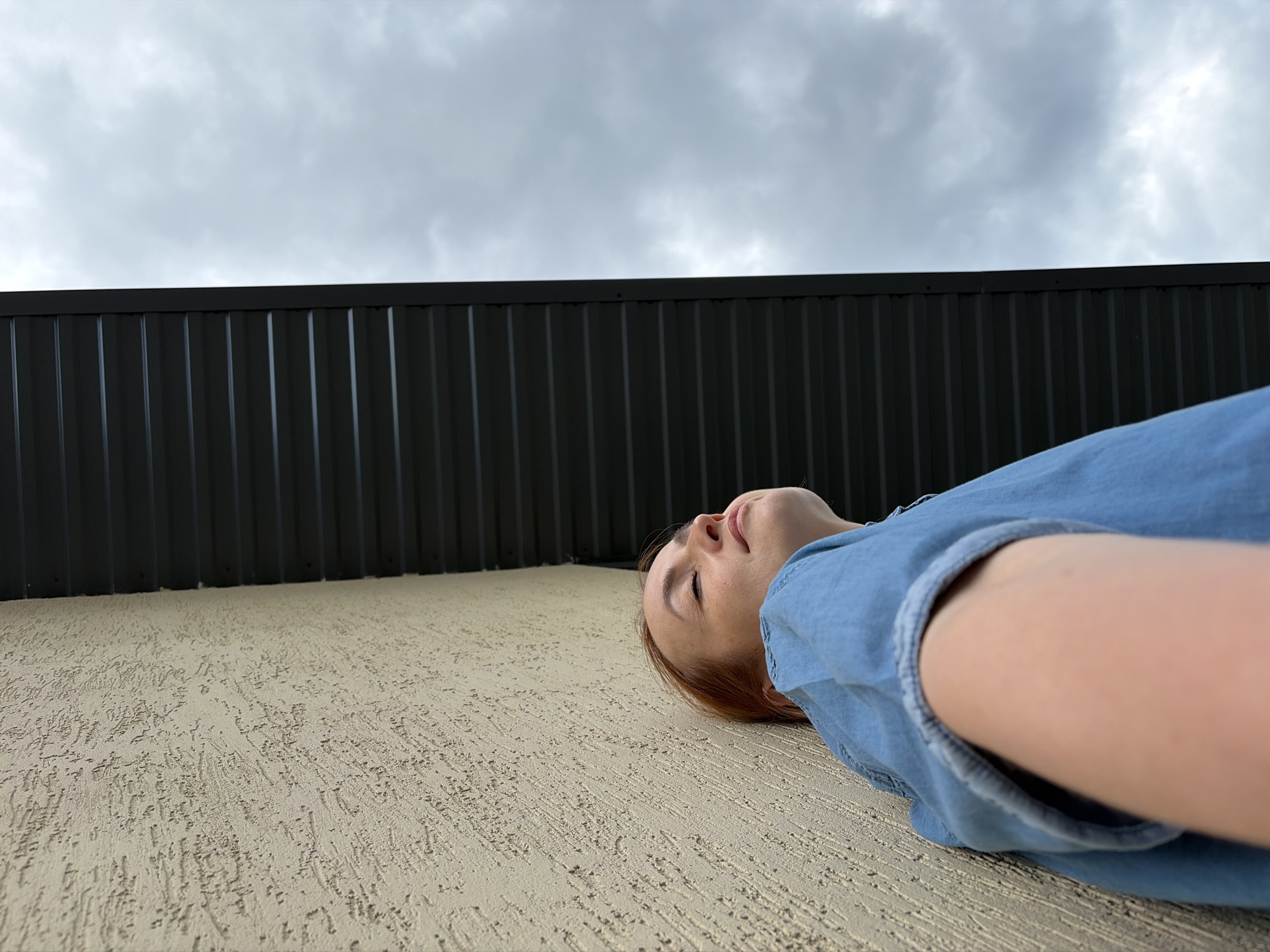 An upward angle shot shows a woman with auburn hair leaning against a textured beige wall, looking upwards with her eyes closed and a serene expression. She is wearing a blue denim shirt. Above and to her right, a dark grey, ribbed roofline runs diagonally across the frame, separating the wall from a cloudy, grey sky. The scene appears to be outdoors, possibly in the city of Chaikovsky, Russia, given the context.
