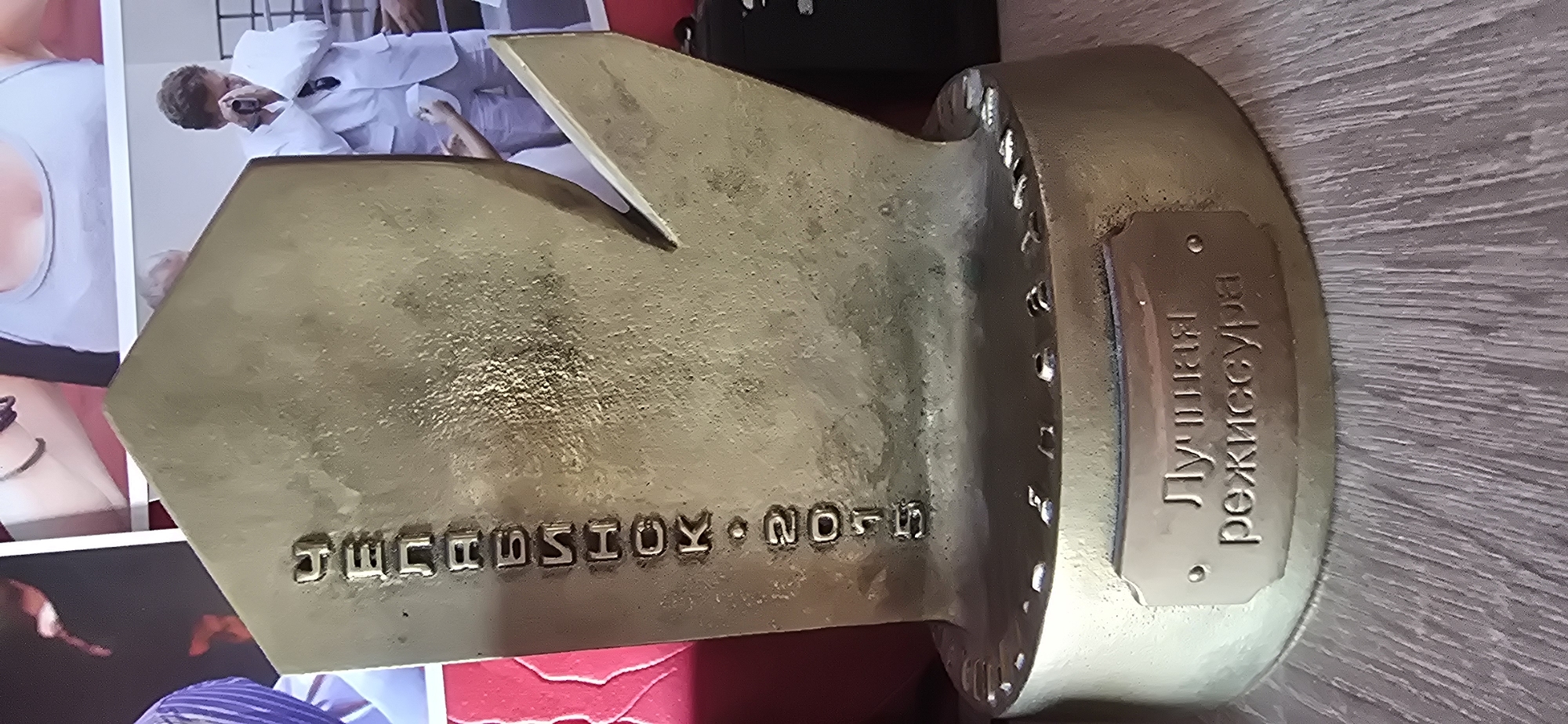 A bronze trophy, likely an award, stands on a wooden surface, possibly a shelf or table. The trophy is predominantly gold-colored with some patina or tarnishing. It features embossed lettering in Russian. The inscription on the side reads vertically: "СЕЗОНА • 2015" which translates to "SEASON • 2015". On the base of the trophy, there is a rectangular plaque with another Russian inscription: "ЛУЧШАЯ РЕЖИССУРА" which translates to "BEST DIRECTION". The trophy has a distinctive shape with a pointed element. In the background, out of focus, are parts of photographs or posters depicting people in various attire, including formal wear and casual clothing. One of the images shows a man in a white suit. The overall environment appears to be indoors, with the wooden surface suggesting a domestic or exhibition setting. The location is identified as Gostagayevskaya, Russia. There are no people directly interacting with the trophy in the frame.