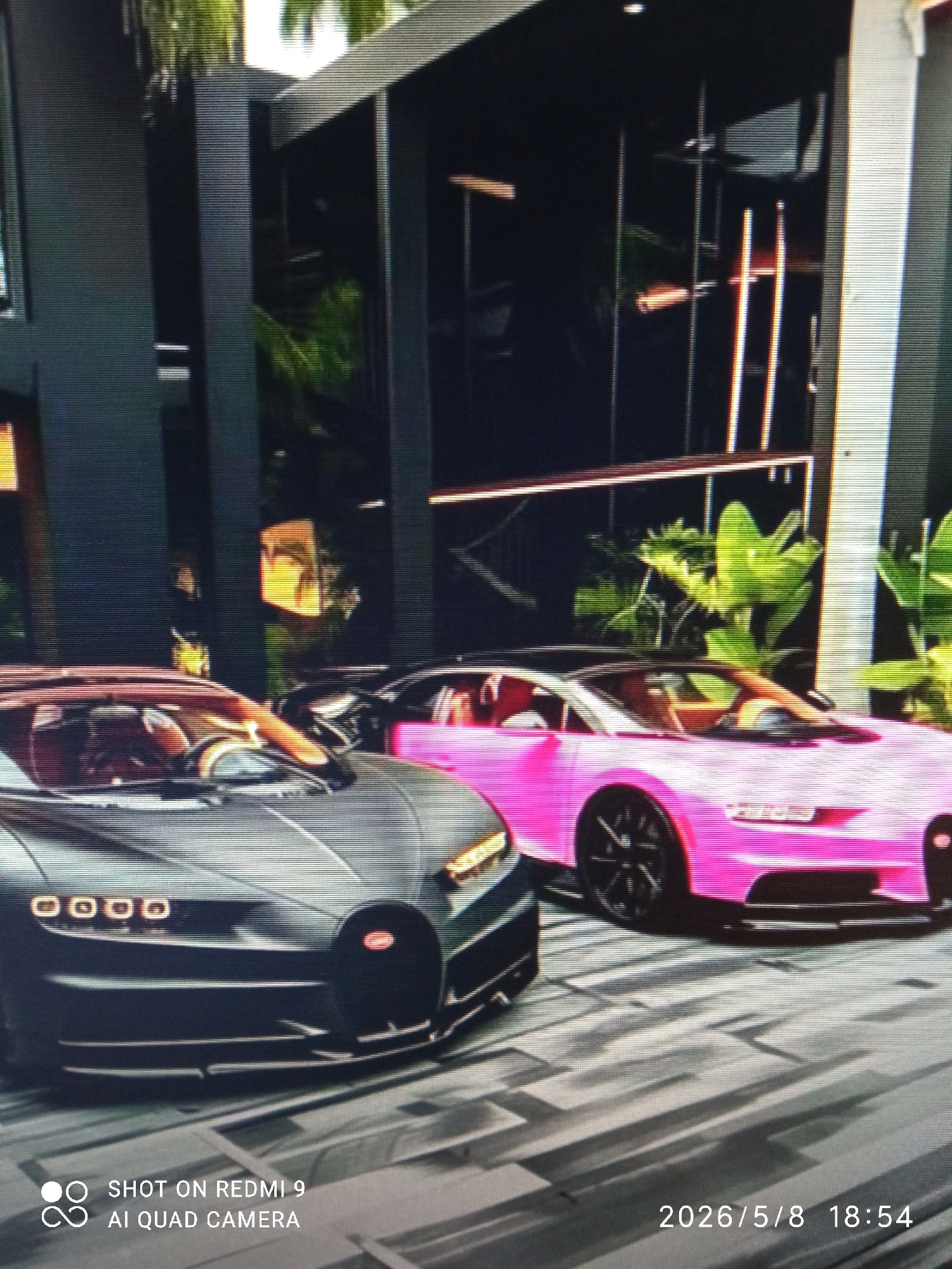 The image shows two Bugatti Chiron supercars parked in front of a modern building. The car on the left is dark grey with an orange interior and the car on the right is pink with a red interior. The building has black columns and large glass windows with plants visible inside. The parking area appears to be paved with grey tiles. The location and city are not identifiable in the image.