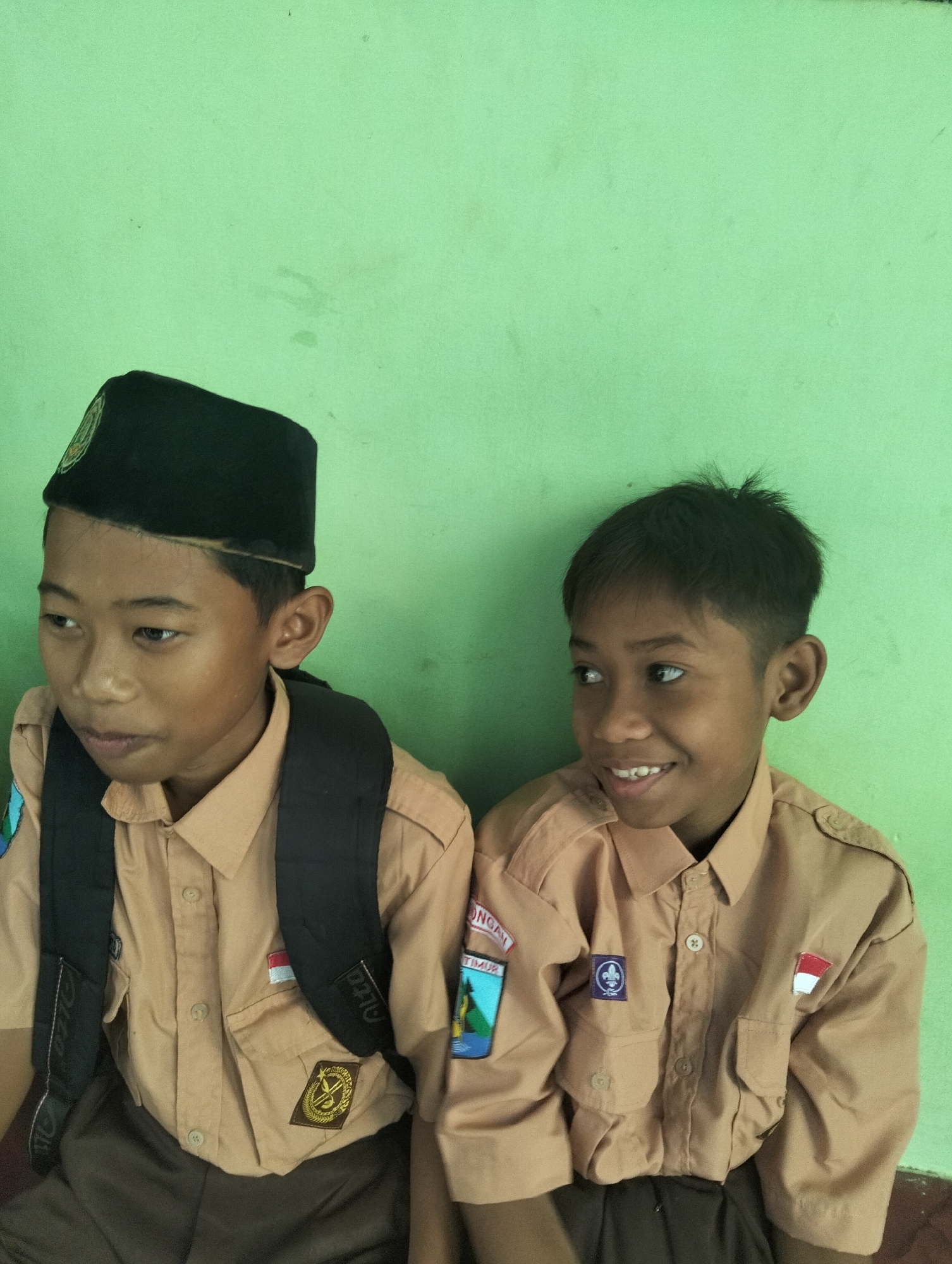 Two young boys, likely of Indonesian nationality given their uniforms, are seated side-by-side against a plain, light green wall. The boy on the left wears a black cap with an emblem and a tan uniform shirt with a dark backpack. He has a backpack with straps visible. The boy on the right wears a similar tan uniform shirt and has short dark hair. Both appear to be students, possibly attending a school in or near Paciran, Indonesia, based on the context provided. The uniforms have patches, including what appears to be a flag of Indonesia on the shirt of the boy on the right. The lighting is soft, suggesting it may be indoors or an overcast day. The wall has some subtle texture and discoloration.