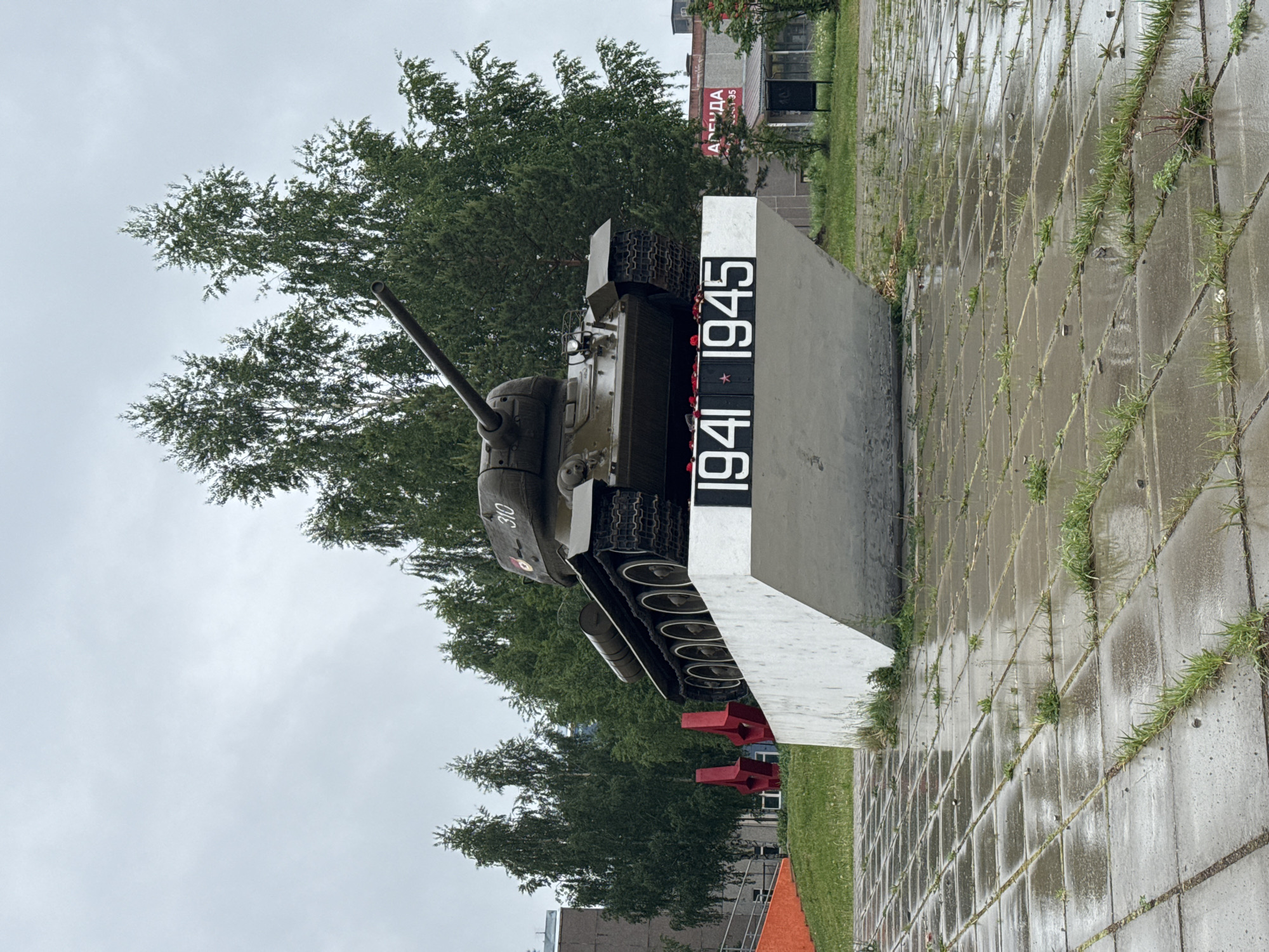 A T-34 tank sits atop a monument commemorating 1941-1945 in Марковский, Russia. The tank is painted green, and the monument is grey and white. The ground is paved with grey tiles with grass growing between the cracks. Trees are behind the tank. The sky is overcast.