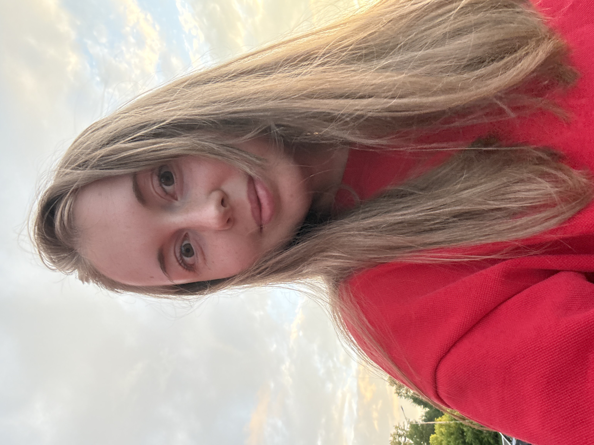 A young woman with long, blonde hair and a red sweatshirt is captured in a close-up selfie against a cloudy sky. Her eyes are open and looking at the camera, and she has a slight smile. The background shows some trees and a lamppost. The lighting suggests it is late afternoon or early evening. The image is taken outdoors, likely in a park or residential area.