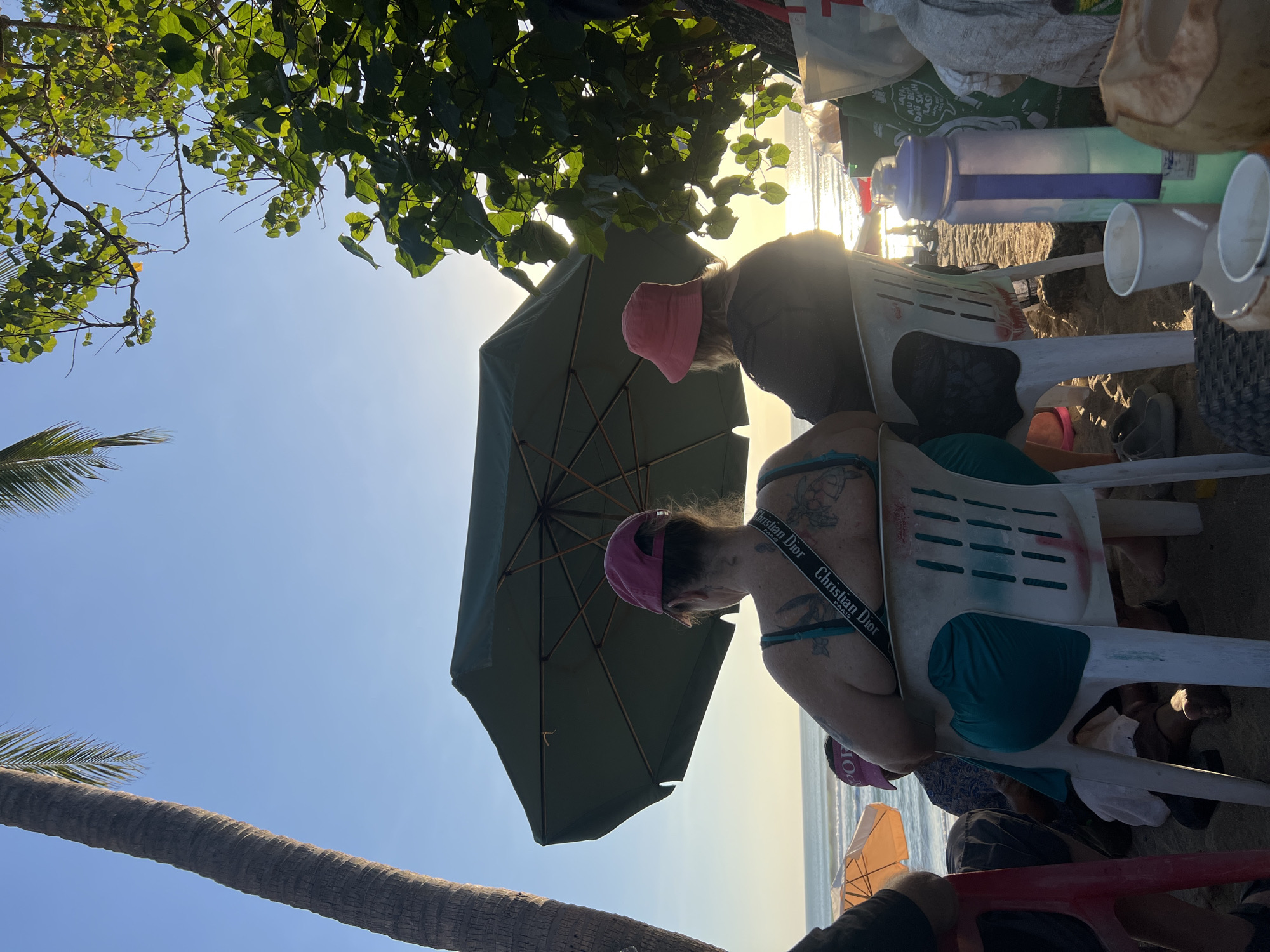 At a beach in Kuta, Indonesia, two individuals are seated on plastic chairs, facing away from the camera. The person on the left wears a pink cap and a teal top, with extensive tattoos visible on their back and arm. A strap with "Christian Dior" branding is slung across their shoulder. The person on the right wears a pink bucket hat and a dark top. They are seated under a large, dark green umbrella, which is angled to provide shade from the late afternoon sun.
The environment is sandy, with the ocean and waves visible in the background. Sunlight glints off the water, indicating it is either sunrise or sunset. To the right of the seated individuals, several objects are visible on a table-like surface, including a large purple and blue water bottle, white paper cups, and a coconut. Palm trees and lush green foliage frame the scene from the left and right, respectively, suggesting a tropical setting. The overall atmosphere appears relaxed, with people enjoying the beach.